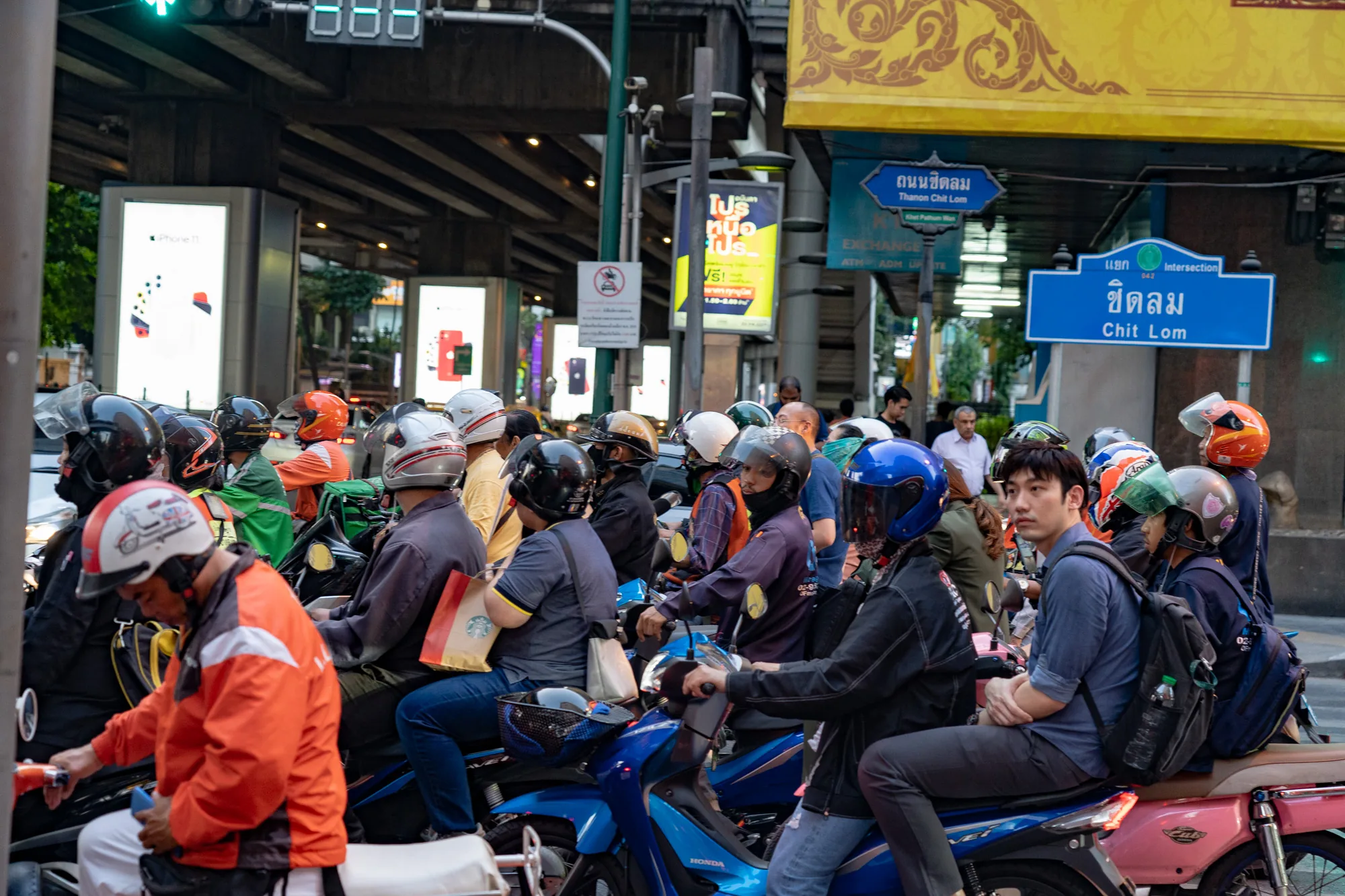 この写真はタイのバンコク、チットロム交差点で撮影されたものです。画面には、信号待ちをしているバイクの群れが写っています。多くの人がヘルメットを着用しており、様々な色やデザインのものが見られます。バイクの色も様々で、青、ピンク、白などがあります。配達員と思われる緑色の制服を着た人もいます。  背景には高架道路と、iPhone 11の広告を含む複数の看板が見えます。道路標識にはタイ語と英語で「Chit Lom」（チットロム）と書かれています。また、「Thanon Chit Lom」（チットロム通り）と書かれた標識も見えます。信号機は青信号を示しており、まもなくバイクの群れが動き出す様子が想像できます。全体的に、活気のあるバンコクの交通風景を捉えた一枚です。