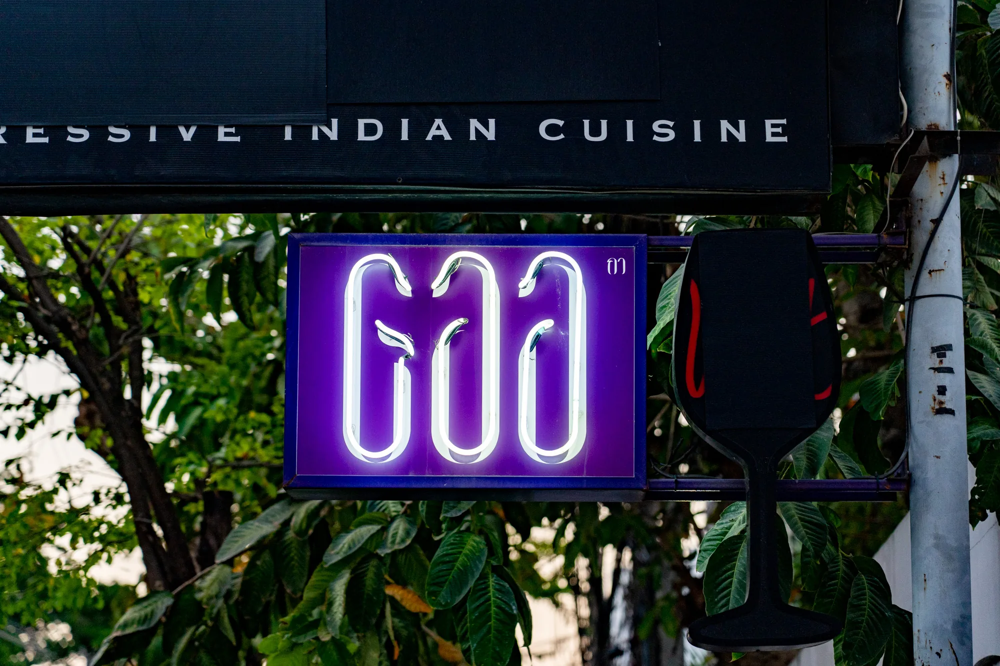 タイのバンコクで撮影された写真には、インド料理店の看板が写っています。看板は二段になっており、上段には「RESSIVE INDIAN CUISINE」と白い文字で書かれています。下段の看板は紫色で縁取られ、ネオンライトでタイ文字と思われるロゴが光っています。その横には小さなタイ文字も添えられています。看板は街路樹の緑に囲まれ、自然光が差し込んでいます。看板の右側にはワイングラスの形をした黒い装飾品が取り付けられ、その横には電柱が立っています。全体的に、街の喧騒を感じさせつつも、スタイリッシュな雰囲気のインド料理店であることが伝わってきます。
