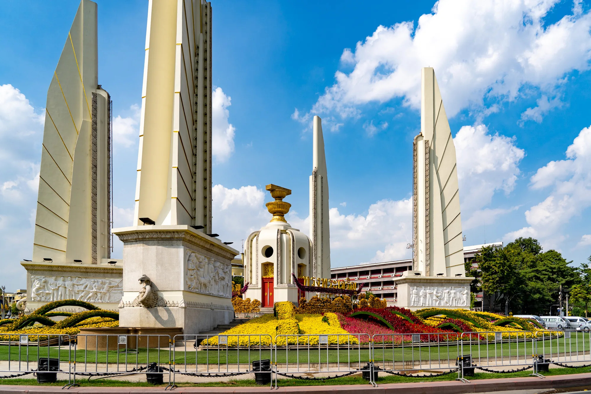 タイのバンコクにある民主記念塔を捉えた写真です。青空に白い雲が浮かび、建造物が明るく照らされています。画面中央には、白い壁に赤いドアが印象的な中心構造物があり、その上に金色の装飾が施されています。構造物の周りには、黄色、赤、紫色の花が丁寧に植えられており、鮮やかな色彩を添えています。記念塔を構成する主要な構造物は、垂直方向に高くそびえ立つ４つの白い柱であり、各々の基部には人物の彫刻が施されています。それらの柱は、中央構造物を取り囲むように配置されています。背景には建物や木々が見え、都会的な風景の一部を形成しています。写真全体から、記念塔の荘厳さと、周囲の美しい景観が伝わってきます。