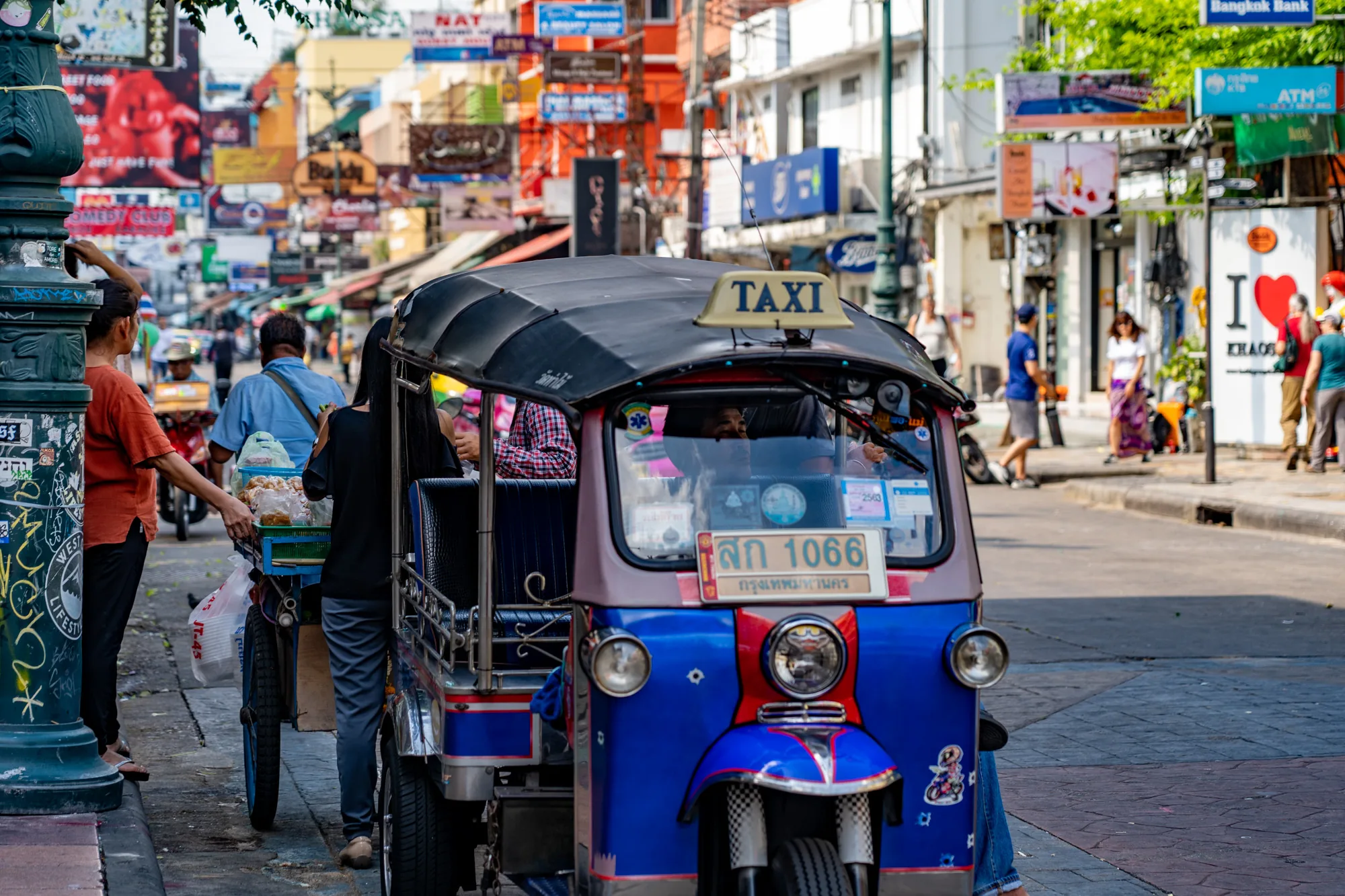 タイのバンコクで撮影された写真です。画面の中央に、鮮やかな青と赤色のトゥクトゥクが大きく写っています。トゥクトゥクは停車しており、運転手や乗客の姿が見えます。ナンバープレートにはタイ語の文字が書かれています。背景には、無数の看板が立ち並び、カオスティックで活気のあるバンコクの街並みが広がっています。看板には英語とタイ語が混在しており、様々な店やサービスを宣伝しているようです。道の端には、食べ物を売る露店商らしき人物がおり、歩行者も何人か見られます。全体的に、東南アジアの都市の喧騒とエネルギーを感じさせる写真です。