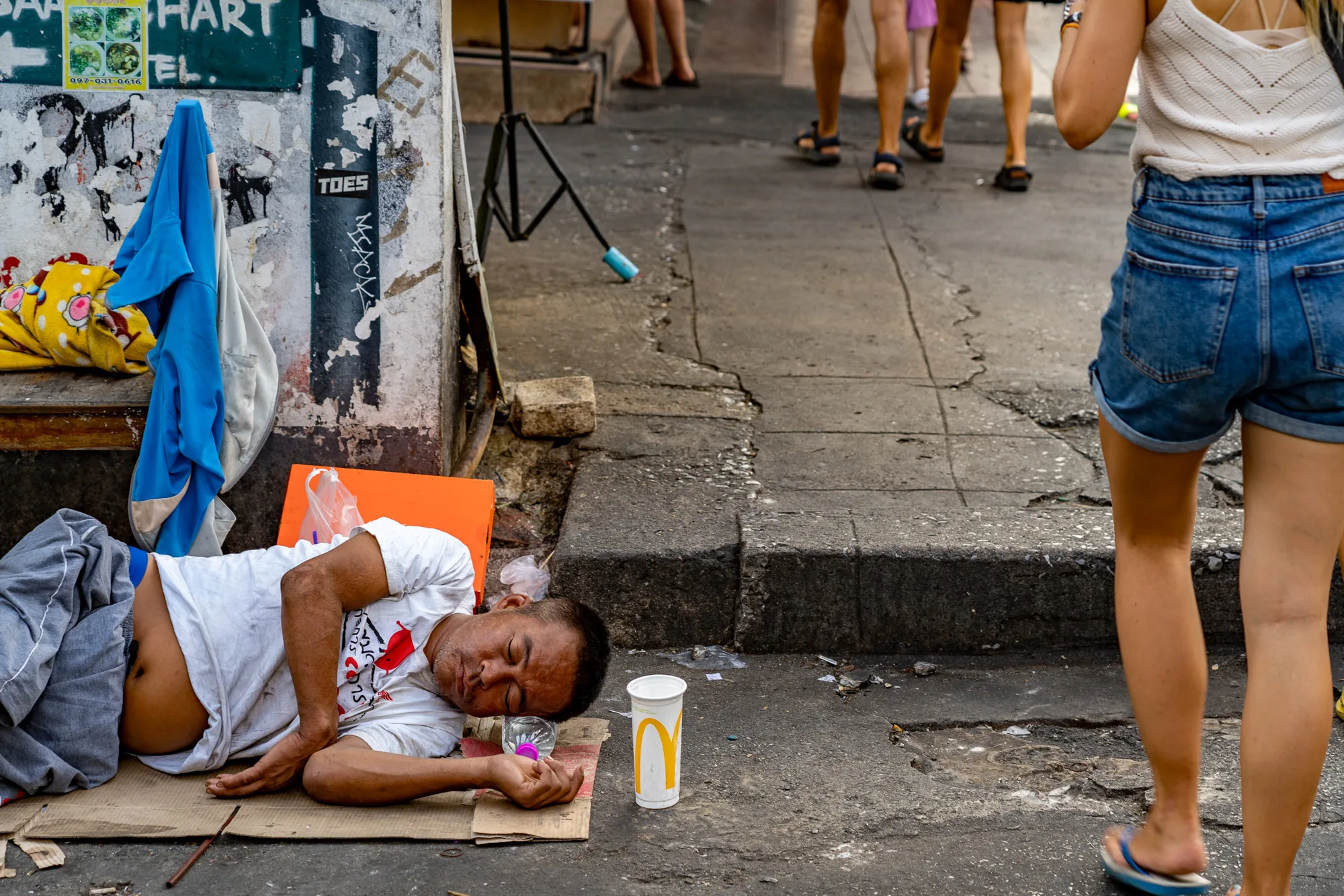 この写真はタイのバンコクで撮影されたもので、路上生活者の男性が写っています。男性は段ボールの上に横たわり、眠っているようです。彼のそばには飲みかけのマクドナルドのカップやペットボトルが置かれています。男性の背景には、落書きや広告が貼られた壁があり、さらに奥には数人の通行人が歩いています。写真全体から、バンコクの都市部の貧困問題や格差が垣間見えます。