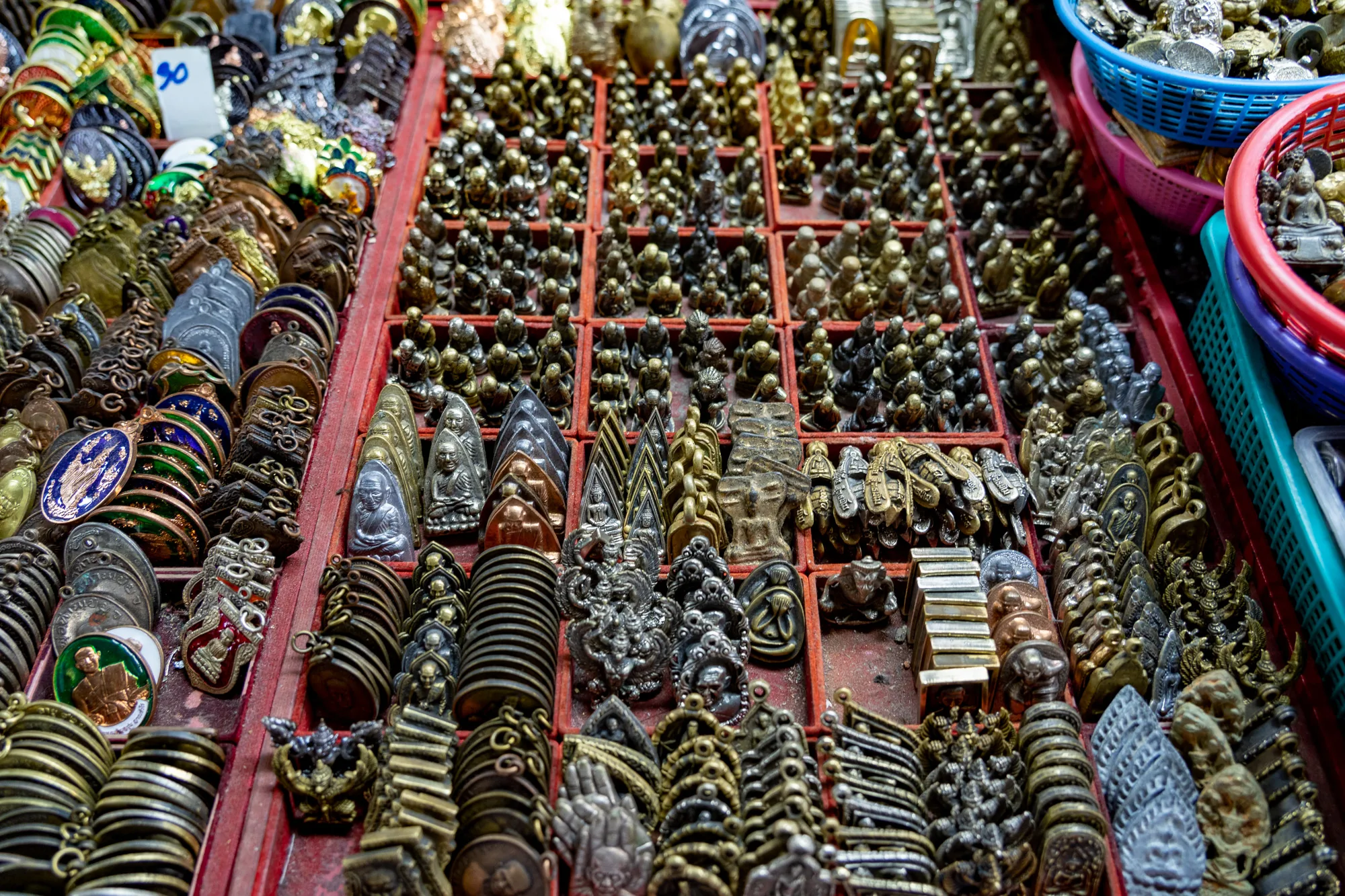 写真は、タイのバンコクの市場で撮影されたお守りの露店を捉えています。赤く仕切られたトレーの中には、様々な種類のお守りが所狭しと並べられています。金、銀、青銅など、多様な金属でできたお守りは、仏像、僧侶の肖像、神話上の生き物など、様々なモチーフをかたどっています。一部のお守りはコイン型で、鮮やかな色で装飾されています。また、小さな仏像が集められたバスケットも写っています。雑多な雰囲気が、活気ある市場の様子を伝えています。お守りのデザインは精巧で、タイの文化や宗教観を反映しているようです。