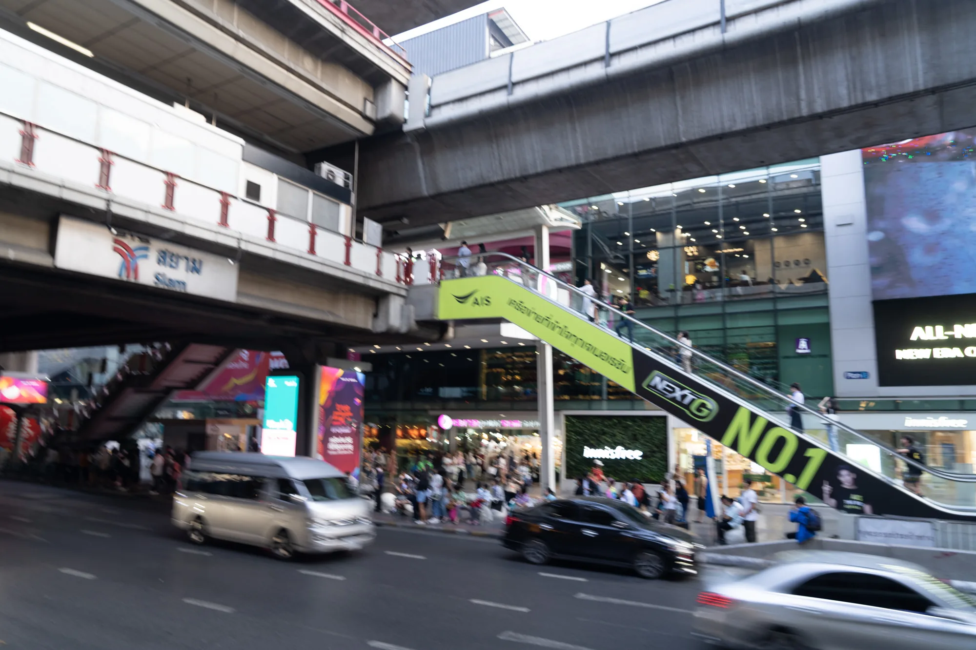 この写真は、タイのバンコクで撮影された街並みのスナップショットです。画面上部には高架鉄道の構造物が見え、「สยาม Siam」と書かれた駅の標識が確認できます。これはおそらく、有名なショッピングエリアであるサイアム駅周辺の風景でしょう。  写真中央には、緑色の看板が付いたエスカレーターが設置されており、その上を人々が移動しています。看板には「AIS」、「NEXTG NO.1」といった文字が見えます。背景には、多くの店舗やレストランが入っているであろう建物が並び、賑やかな雰囲気が伝わってきます。  画面下部には道路が写っており、バン、黒いセダン、そして白い車の動きが捉えられています。人通りも多く、活気のある都会の様子が伝わってきます。全体的にややブレた描写から、移動中に撮影された写真であると考えられます。