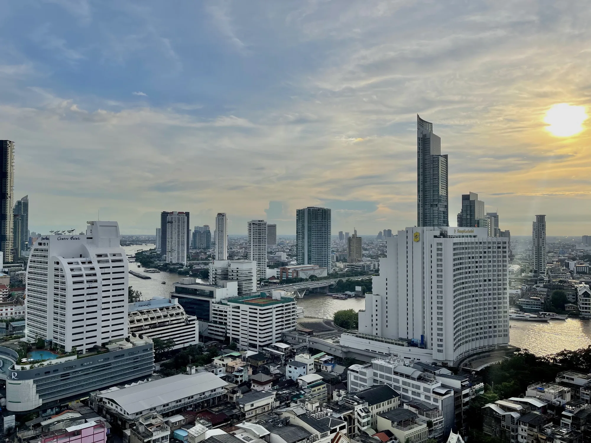 この写真は、タイのバンコクにおける都市の景観を捉えています。高所から撮影されたと思われるこの画像には、チャオプラヤー川が流れ、その両岸に高層ビル群が立ち並ぶ様子が広がっています。空には薄い雲が広がり、太陽が柔らかい光を都市に降り注いでいます。建物の多くは白または淡い色で塗られており、整然とした印象を与えます。川には船が航行し、橋が架かっています。手前には、密集した低層の建物が建ち並び、その屋根の形状や色彩が多様で、都市の複雑さを感じさせます。全体的に、都市の発展と伝統的な生活が共存するバンコクの独特な雰囲気が表現されている写真です。