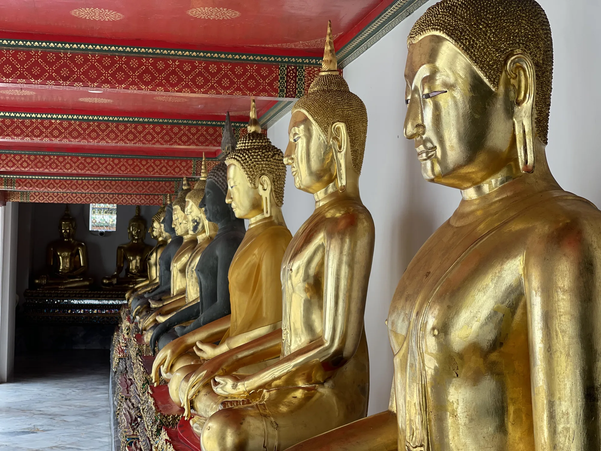 写真はタイのバンコクで撮影されたもので、寺院の一角に並んだ仏像群を写しています。仏像は全て座禅を組んだ姿勢で、正面を向いています。手前の仏像は金色に輝いており、光の反射が美しいです。奥に続くにつれて、いくつかの仏像は黒色をしています。天井は赤色で、金色の模様が描かれており、タイの伝統的な装飾が施されています。全体的に落ち着いた雰囲気で、神聖な空間が表現されています。仏像の表情は穏やかで、見る人に安らぎを与えるようです。