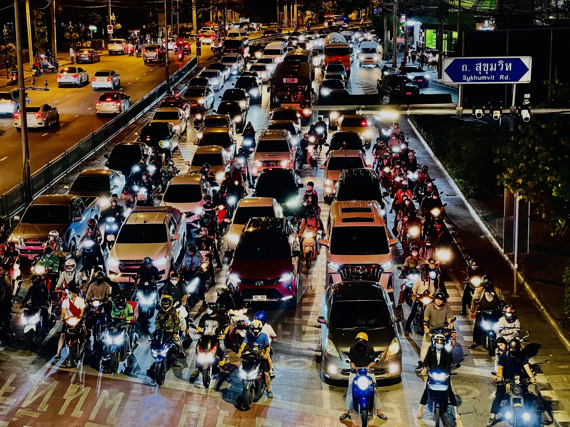 この写真はタイのバンコクで夜に撮影されたもので、交通渋滞の様子を捉えています。道路は車とバイクで埋め尽くされており、特にバイクの数が非常に多いのが特徴的です。車のヘッドライトや街灯が夜の街を照らし、活気のある雰囲気を出しています。スクンビット通り(Sukhumvit Rd.)への標識が見え、場所を特定できます。多くの人がマスクを着用しており、コロナ禍における都市の様子が伺えます。