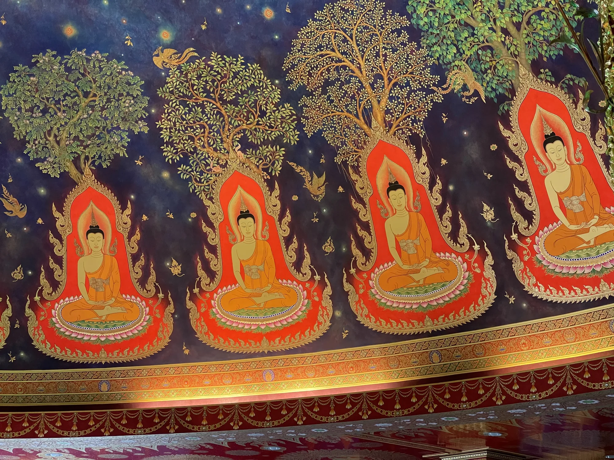 これはタイのバンコクで撮影された寺院の天井画です。画面全体にわたって、4体の仏陀像が描かれています。それぞれの像は蓮の花の上に座禅を組んでおり、後ろには燃え盛る光輪のような装飾が施されています。各仏陀の上には、緑豊かな木が描かれており、空は紺色で星が散りばめられています。木の間には、金色の鳥が飛んでいます。絵全体は鮮やかな色彩で、特にオレンジ、赤、金色が目立ち、タイの伝統的な芸術様式を示しています。画像の下部には、装飾的な縁取りが見られ、天井全体が非常に精巧に装飾されていることがわかります。
