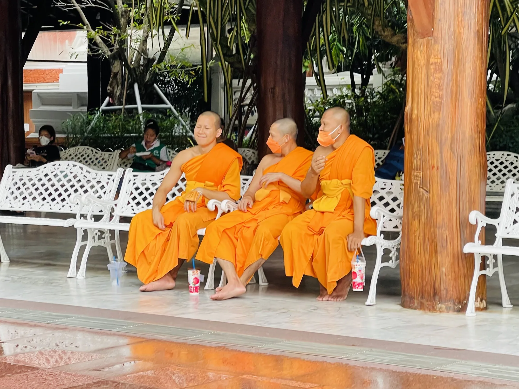 写真はタイのバンコクで撮影されたもので、オレンジ色の袈裟を身につけた3人の僧侶が、白い装飾が施されたベンチに座って休憩している様子を捉えています。僧侶たちはマスクを着用しており、新型コロナウイルスの感染対策を意識していることが伺えます。それぞれ飲み物を持っていたり、裸足であったりと、リラックスした様子です。背景には緑豊かな植物や寺院のような建造物が見え、穏やかな雰囲気が漂っています。他の人たちもベンチに座って休憩しており、地元の人々の日常風景の一コマを切り取ったような写真です。