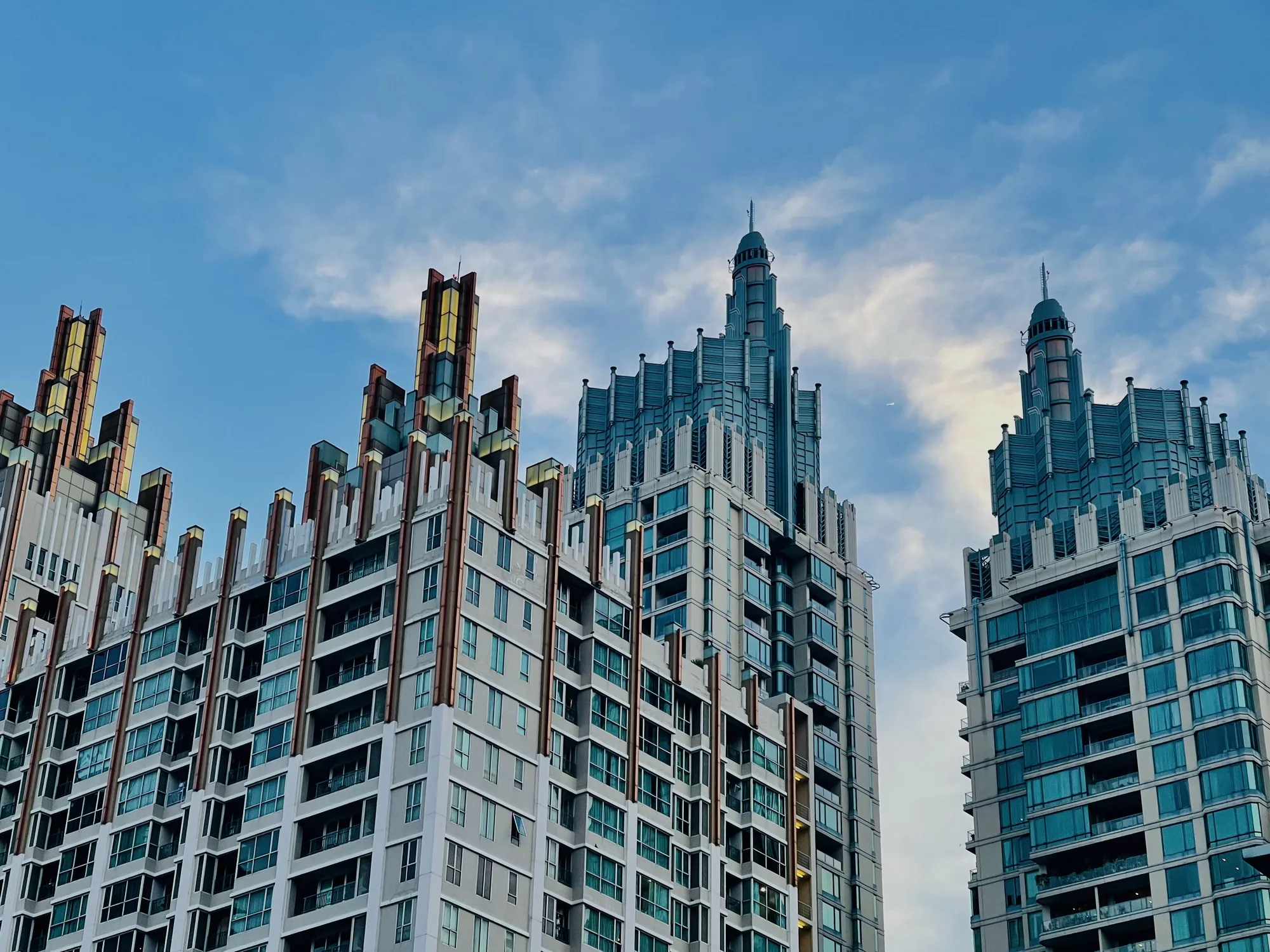 写真には、タイのバンコクにある高層ビル群が青空を背景に写っています。画面には3棟のビルが主要な被写体として捉えられており、それぞれ異なるデザインが施されています。一番左のビルは、頂部に茶色と黄色の装飾が施された複数の柱が特徴的です。中央のビルは、段々になったデザインで、銀色の装飾が施されています。右側のビルも同様に段々としたデザインですが、こちらは青みがかった銀色で統一されています。各ビルには多数の窓があり、高層マンションまたはオフィスビルであると思われます。空は薄い雲が浮かぶ青空で、建物の輪郭を際立たせています。全体的に、近代的な都市景観を捉えた写真であり、建築デザインの多様性と都市の発展を感じさせます。