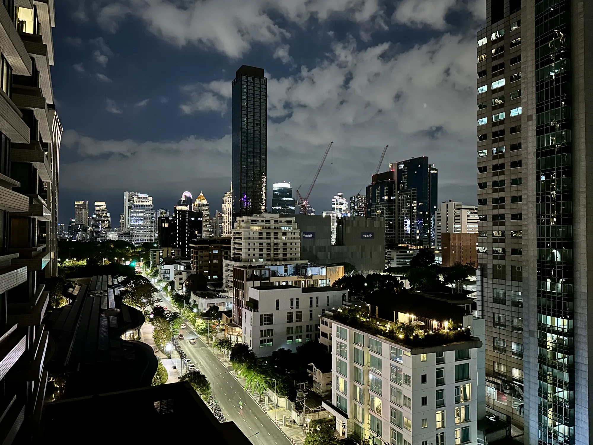 この写真はタイのバンコクで夜に撮影された都市の風景です。高い位置から撮影されたようで、手前には建物の一部が写り込んでおり、その向こうには高層ビル群が立ち並んでいます。空には雲が広がり、街の明かりがそれらをわずかに照らしています。いくつかの高層ビルは建設中で、クレーンが確認できます。道路は街灯で照らされ、交通量は比較的少ないようです。屋上庭園がある建物も見られます。全体的に、都会の夜の静けさと発展のエネルギーが感じられる雰囲気です。
