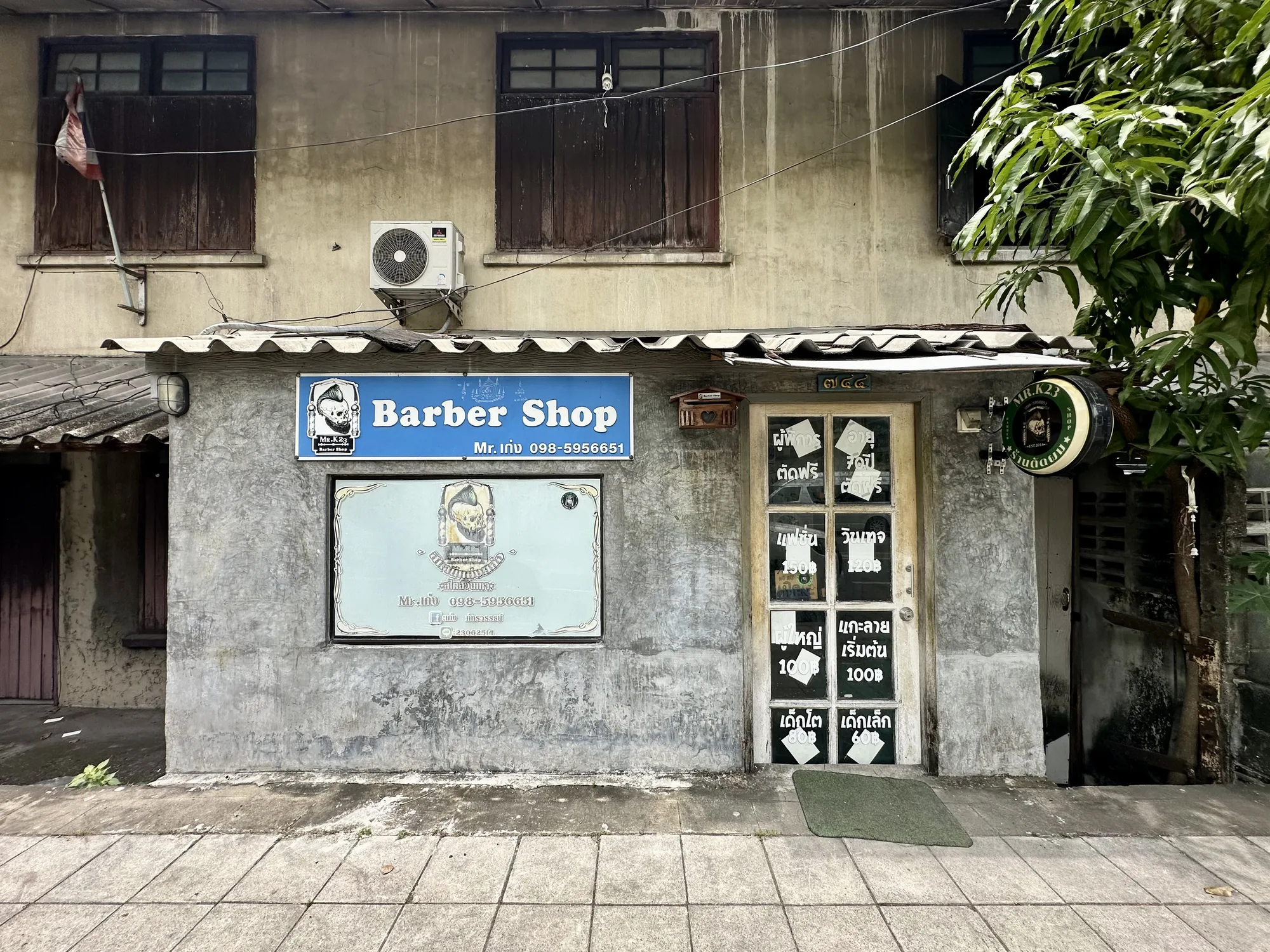 写真はタイのバンコクで撮影された、路地に面した小さな床屋の外観を捉えています。建物はコンクリート造りで、壁は灰色で、ところどころ汚れや劣化が見られます。建物の上部には木製の窓が3つあり、窓枠は濃い茶色で、古びた様子です。左端の窓にはタイの国旗が掲げられています。窓の下にはエアコンの室外機が設置されています。  床屋の入り口はガラスのドアになっており、ドアにはタイ語の料金表が手書きで書かれた紙が貼られています。ドアの前には緑色のマットが敷かれています。ドアの横には円形の看板があり、店名とロゴが描かれています。看板の上には小さなメールボックスのようなものが取り付けられています。  写真全体を通して、下町の雰囲気が漂っており、生活感のある様子が伺えます。右側には緑豊かな植物が写っており、都会の中の自然を感じさせます。