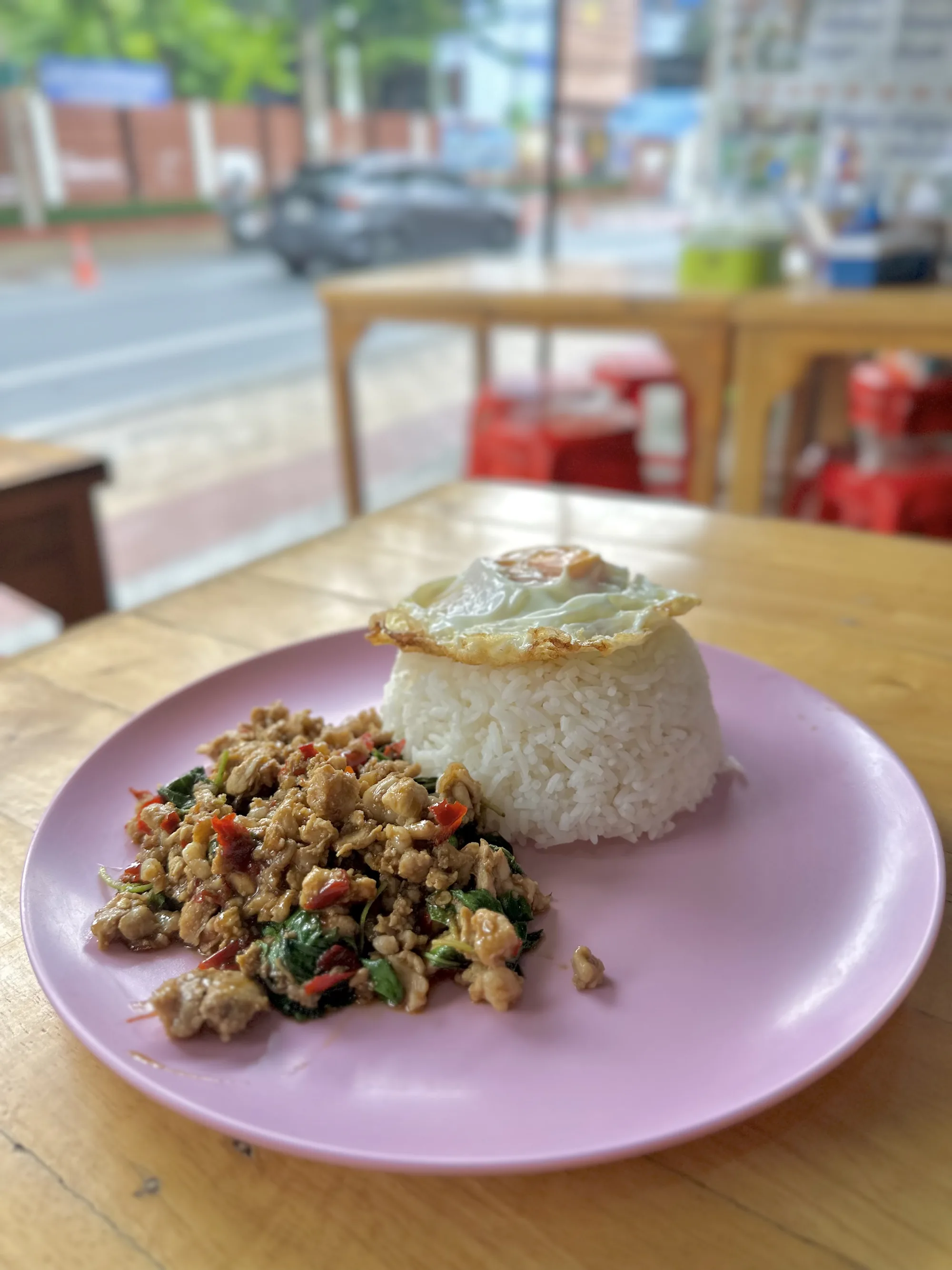 写真は、タイのバンコクで撮影されたと思われるガパオライスの料理写真です。手前のテーブルには、ピンク色の皿に盛られたガパオライスが置かれています。ご飯はこんもりと丸く盛られ、その上には目玉焼きがトッピングされています。ガパオは鶏ひき肉が使われており、バジルや唐辛子なども一緒に炒められているようです。全体的に美味しそうな照りがあり、食欲をそそります。背景には、ぼやけていますが、道路や建物が見え、お店のテラス席のような場所で撮影されたことが伺えます。テーブルや椅子の色合いも温かく、リラックスした雰囲気です。