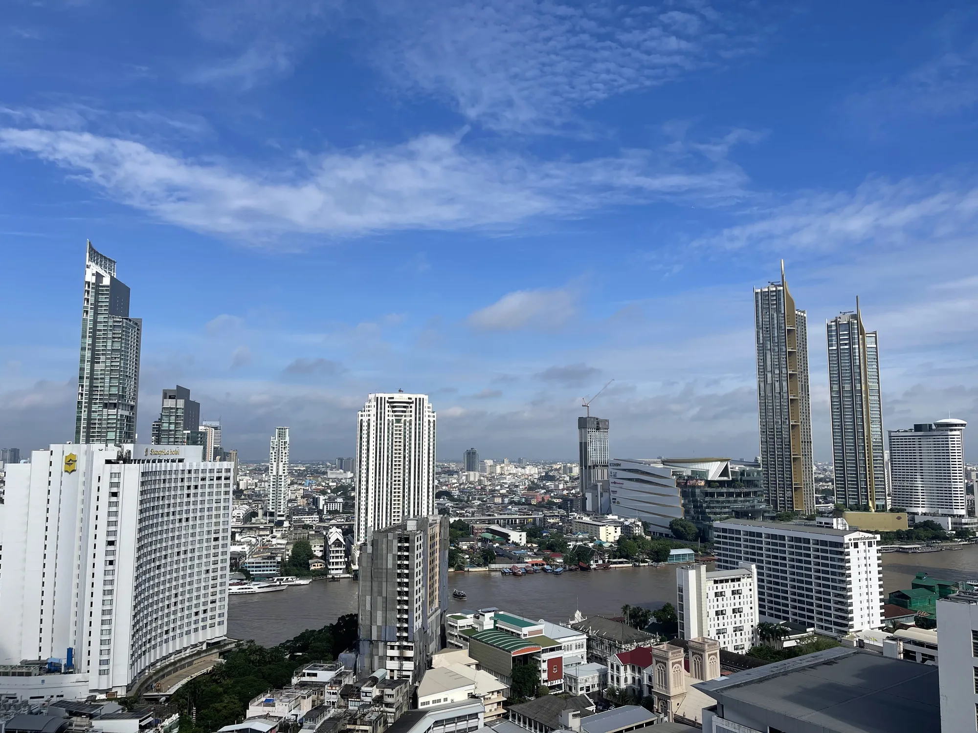 写真はタイのバンコクで撮影されたもので、高層ビルが立ち並ぶ都市景観を捉えています。画面中央にはチャオプラヤー川が流れ、その両岸には建物が密集しています。空は青く、白い雲が点在しており、晴れた日の撮影であることが分かります。写真全体として、活気がありながらも穏やかなバンコクの雰囲気を伝えています。画面左手前にはシャングリ・ラ ホテルがあり、右側にはアイコンサイアムや建設中の高層ビルが見えます。川にはいくつかのボートが航行しており、都市の喧騒と自然の調和を感じさせます。