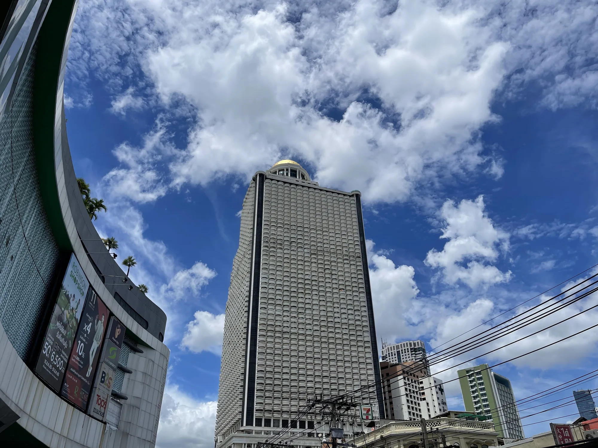 タイのバンコクで撮影された写真です。画面中央には、細長いバルコニーが多数並んだ独特なデザインの高層ビルがそびえ立っています。ビルの頂上には金色のドーム状の構造物があり、目を引きます。空は青く、白い雲が点在しており、建物を背景に美しいコントラストを生み出しています。  画面左側には、一部が切り取られた現代的な建物の一部が見え、看板には英語とタイ語の文字が確認できます。電線が画面を横切っており、都市の風景の一部となっています。写真全体を通して、晴天の日のバンコクの賑やかな雰囲気が伝わってきます。