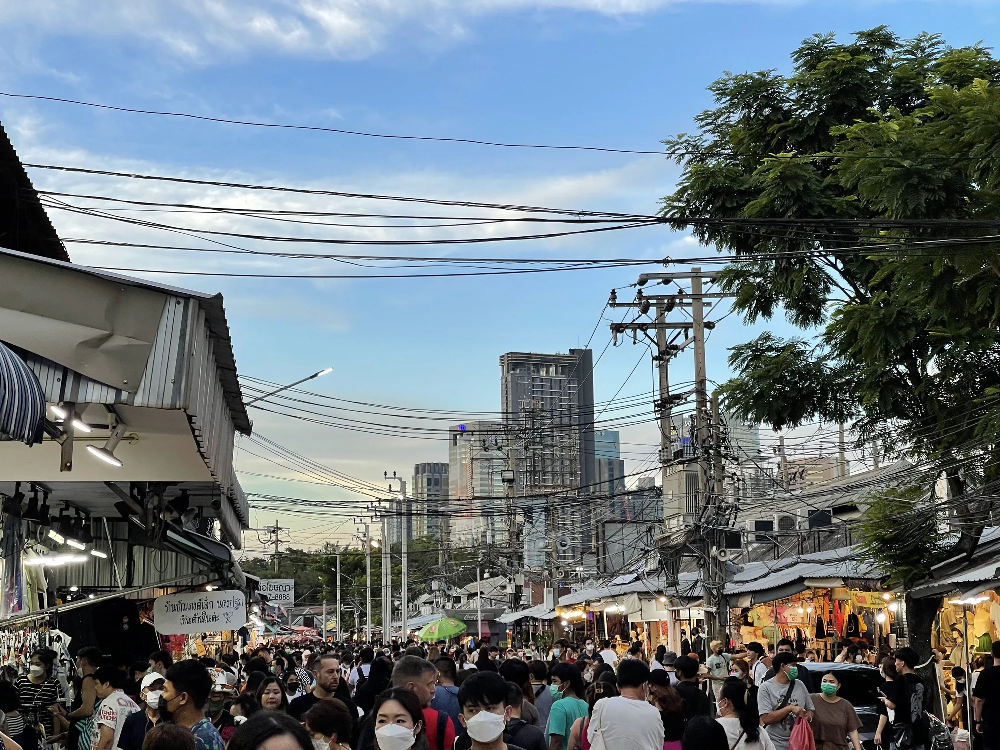 この写真はタイのバンコクで撮影された、賑やかな市場の様子を捉えています。画面いっぱいに人がひしめき合っており、多くがマスクを着用しています。画面奥には高層ビル群が立ち並び、都市部の風景が広がっています。電線が複雑に張り巡らされ、アジアらしい雑多な雰囲気を醸し出しています。両側には屋台が並び、衣類や雑貨などが販売されているようです。看板にはタイ語の文字が見えます。空は薄い雲が広がる夕暮れ時でしょうか、少しオレンジがかった光が全体を照らしています。活気と混沌が入り混じった、バンコクのリアルな日常が伝わってくる一枚です。
