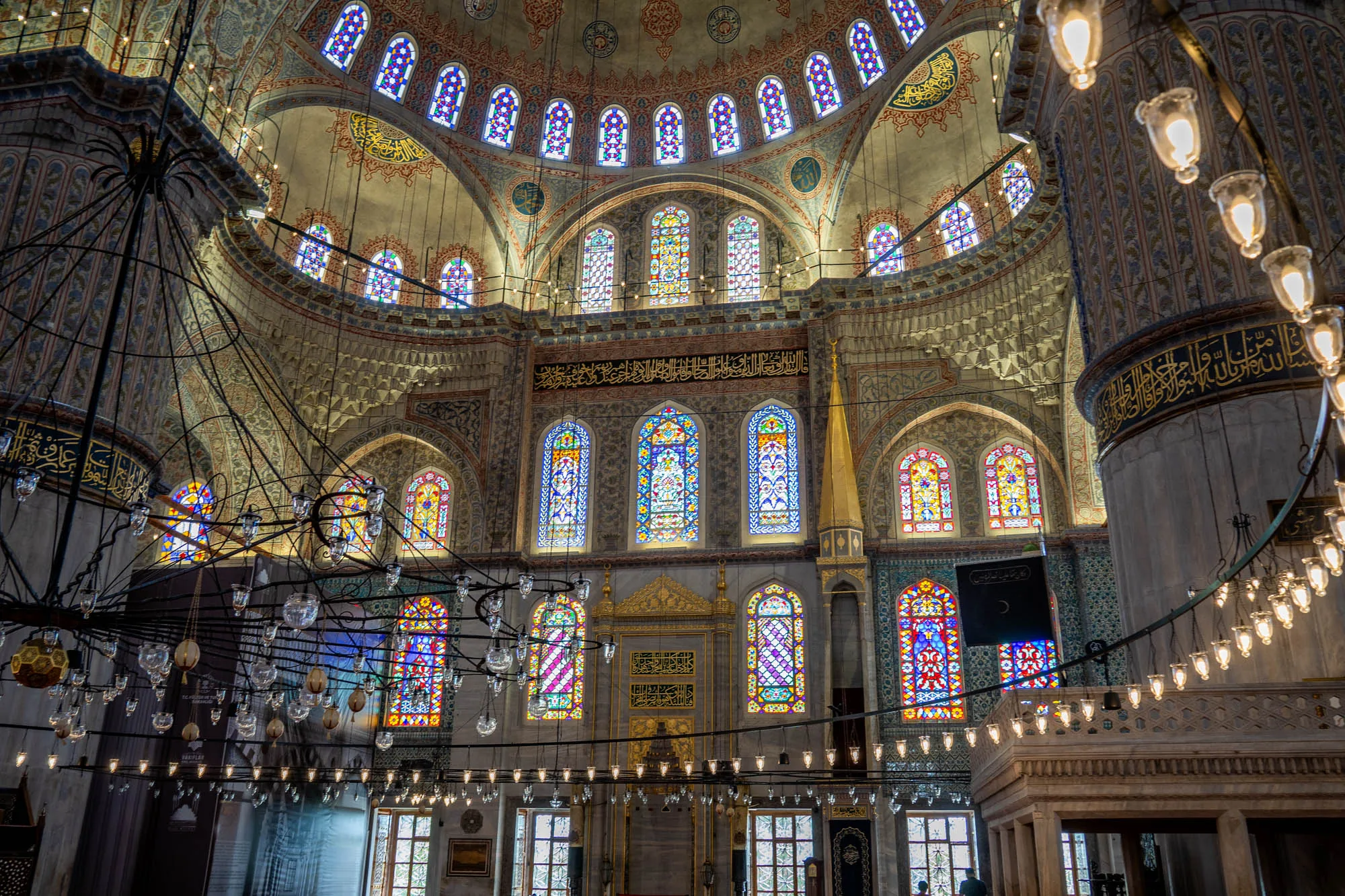 写真はトルコのイスタンブールにあるモスクの内部を捉えたものです。天井はドーム型で、美しい模様と装飾が施されています。ステンドグラスの窓が多数あり、そこから差し込む光がモスク内を幻想的に照らしています。巨大なシャンデリアが吊り下げられており、その装飾的なデザインが印象的です。壁面にはアラビア文字が描かれ、イスラムの雰囲気を醸し出しています。全体的に荘厳で静謐な雰囲気が感じられます。