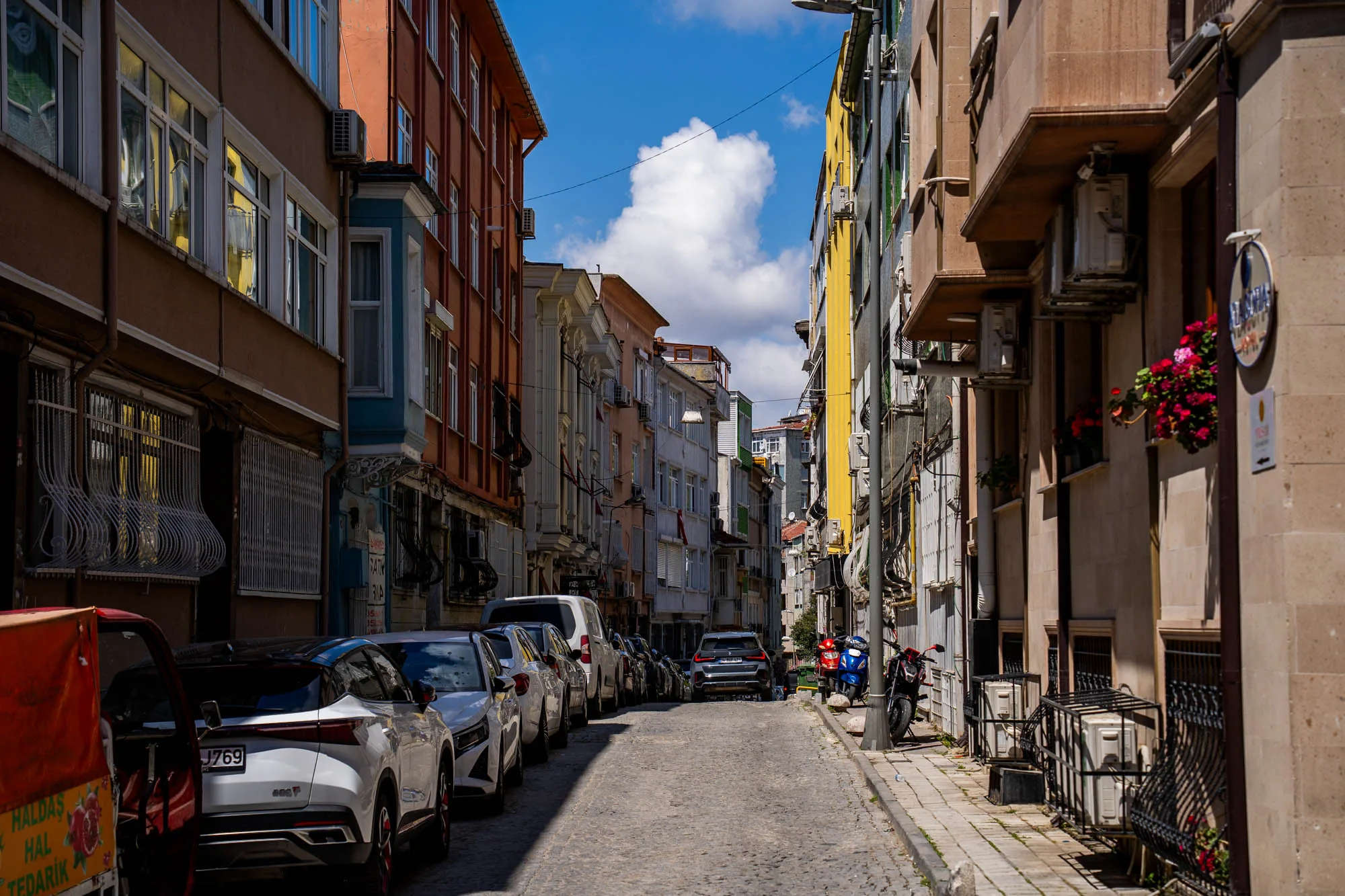 イスタンブールの石畳の道が続く街並みの写真です。道の両側には様々な色の建物が立ち並び、空には白い雲が浮かんでいます。建物はオレンジ、黄色、ベージュなどカラフルで、それぞれ異なるデザインの窓や装飾が施されています。多くの建物にはエアコンの室外機が設置されています。道の左側には車が縦列駐車しており、右側にはオートバイが停まっています。歩道には花が飾られた窓辺があり、生活感を感じさせます。全体的に落ち着いた雰囲気ですが、建物の色彩が豊かで、独特の魅力があります。