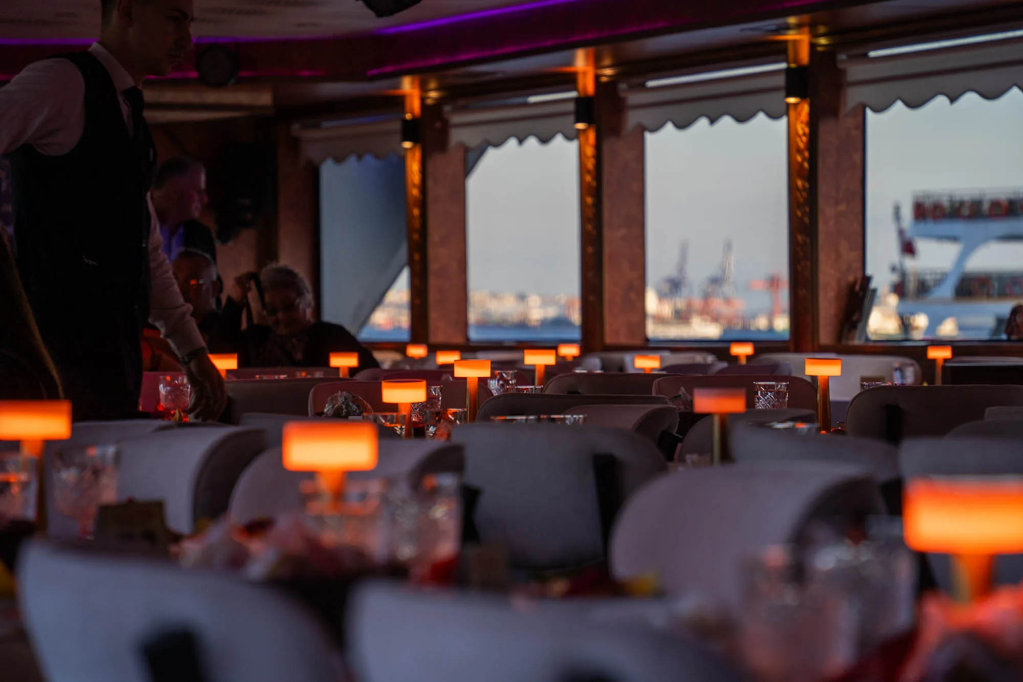 この写真はトルコのイスタンブールで撮影されたレストランの様子を捉えています。レストランはボートの中にあるようで、窓からは対岸の景色が見えます。テーブルには小さなオレンジ色の照明が置かれており、それが室内を暖かく照らしています。ウェーターが準備をしている様子や、食事をしている客の姿も見られます。全体的に落ち着いた雰囲気で、夜景を楽しみながら食事をするのに最適な場所のようです。
