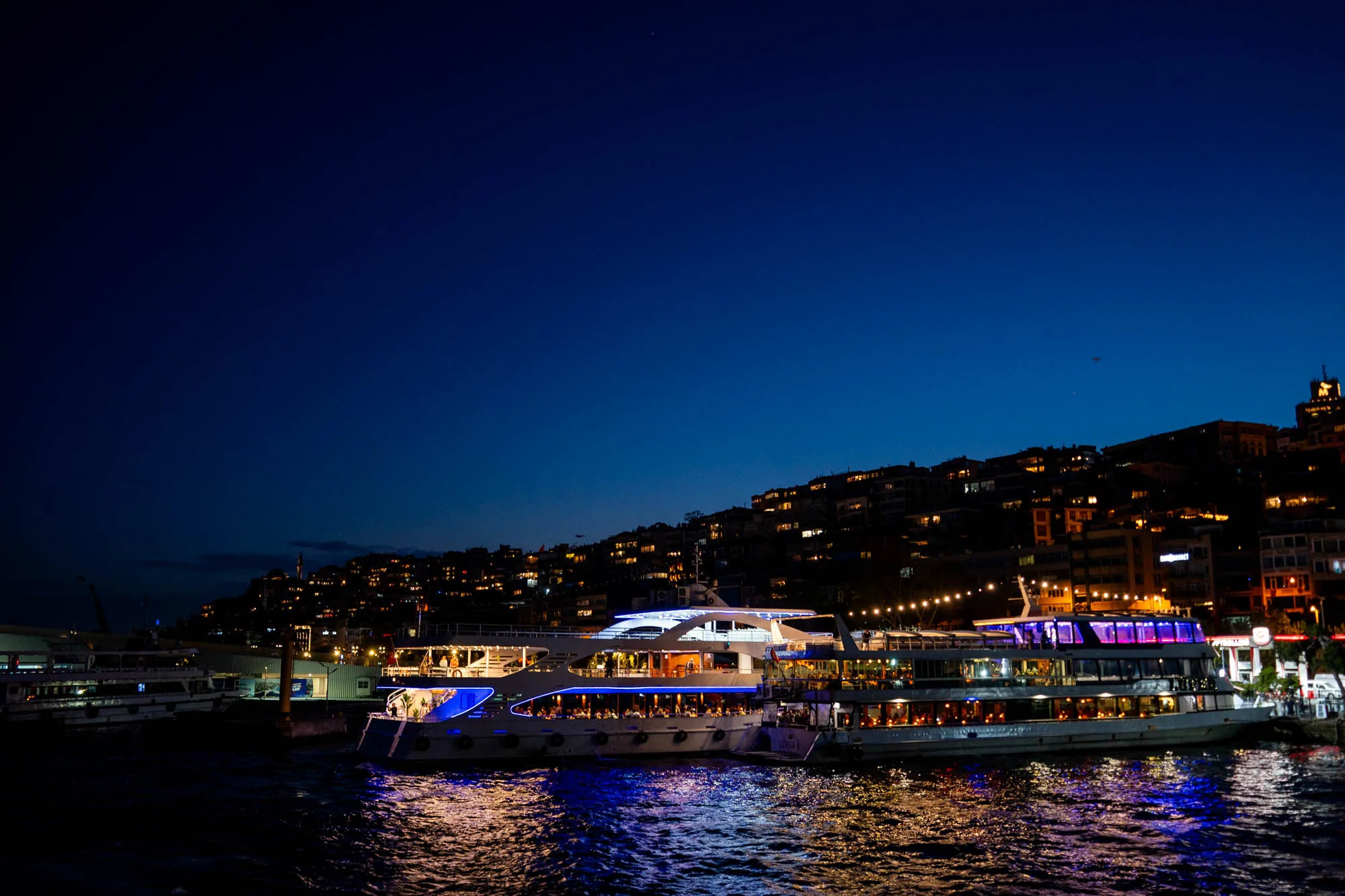 トルコのイスタンブールで撮影された夜景の写真です。深い青色の空の下、水面に浮かぶ複数の船が主な被写体となっています。これらの船はレストラン船のようで、窓からは明るい光が漏れ、内部に人々がいる様子が伺えます。船の縁には青いライトが装飾されており、水面に反射して美しい光の模様を作り出しています。背景には、丘の斜面に沿って建てられた建物群が見え、建物からも暖かい光が灯っています。全体的に、ロマンチックで賑やかな雰囲気を醸し出しており、夜のイスタンブールの魅力を伝えています。