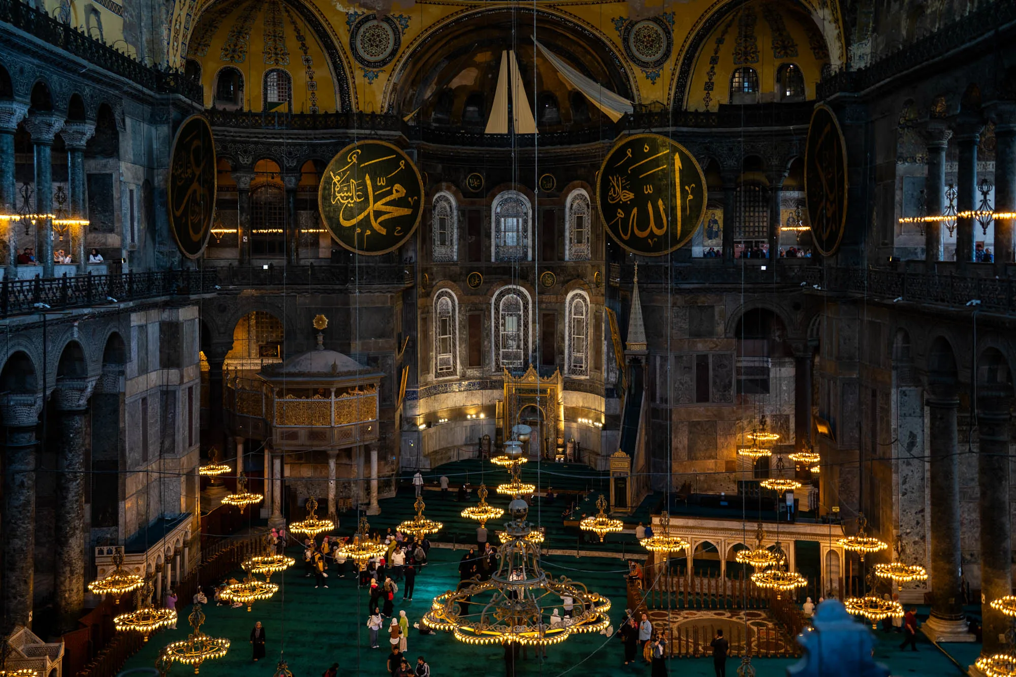 この写真は、トルコのイスタンブールにある寺院の内部を捉えたものです。高い天井には装飾が施され、巨大なアラビア文字が書かれた円盤が吊るされています。内部は緑色のカーペットで覆われ、多くの人々が訪れています。豪華なシャンデリアが並び、温かい光を放ち、荘厳な雰囲気を醸し出しています。石造りの壁や柱が歴史を感じさせ、観光客がその美しさを堪能している様子が伺えます。