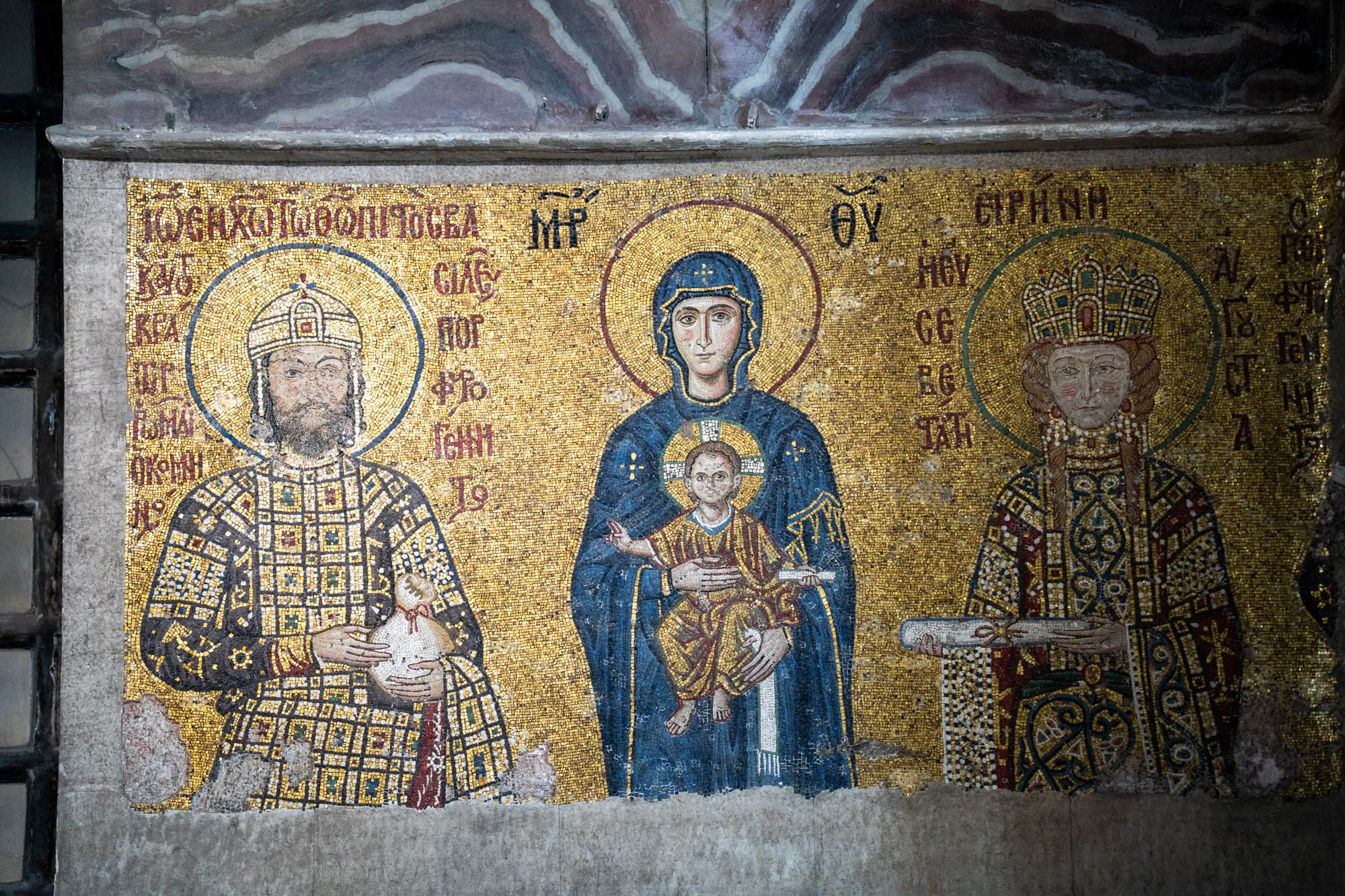 この写真はトルコのイスタンブールで撮影されたモザイク画です。金色の背景に、左から順に、男性、聖母マリアと幼子イエス、そして女性が描かれています。男性はひげを生やし、豪華な模様の服を着て、頭には光輪が描かれています。聖母マリアは青い服を着ており、幼いイエスを抱いています。幼子イエスにも光輪があります。右側の女性も豪華な服と王冠を身に着け、光輪があります。モザイクは細かいタイルで構成されており、古さを感じさせます。全体的に厳かで荘厳な雰囲気を醸し出しています。