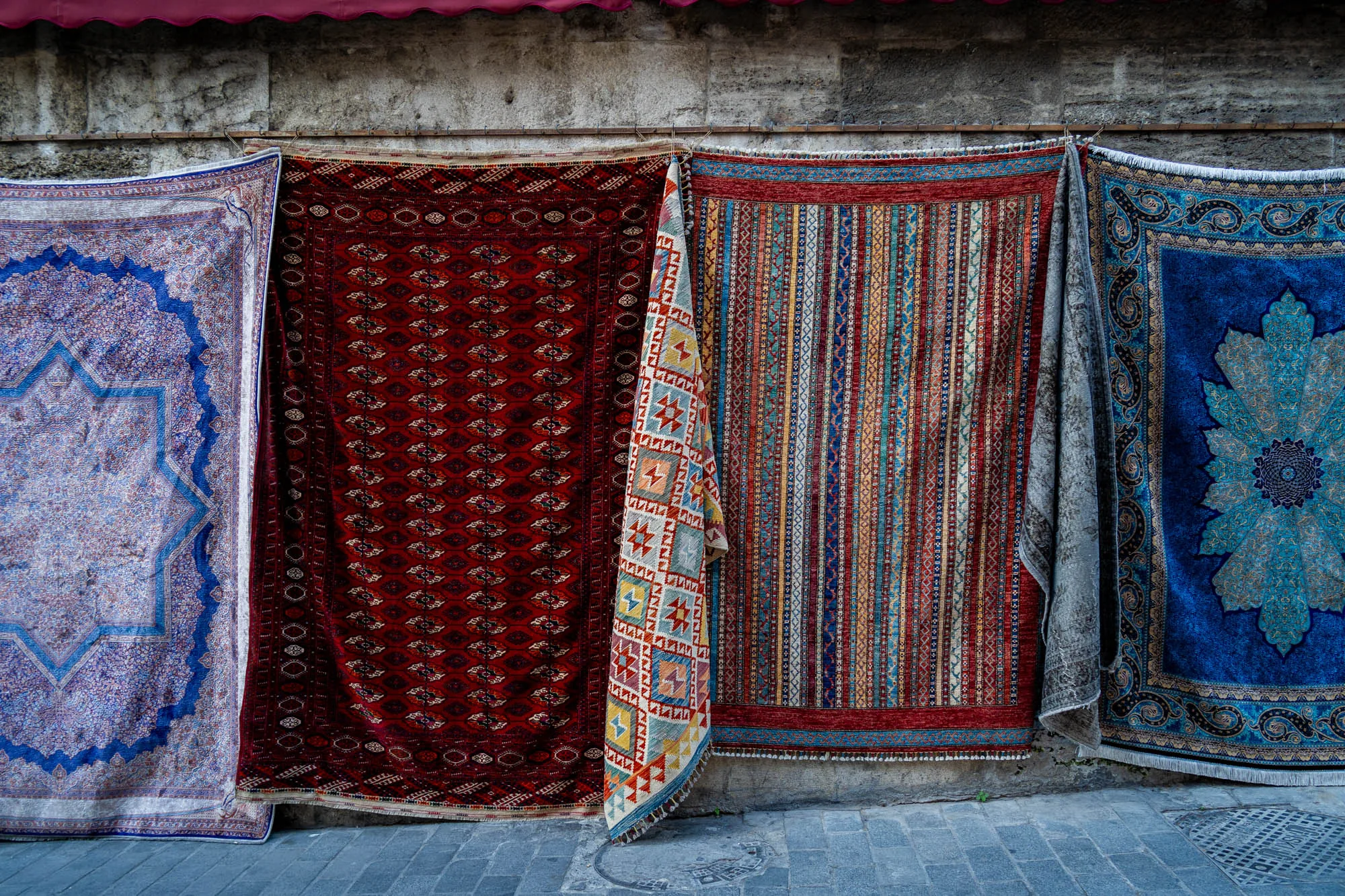 写真には、トルコのイスタンブールにある店の外壁に掛けられた様々な種類の絨毯が写っています。壁は石造りで、少し古びた雰囲気があります。壁の上部には赤い日よけが見えます。絨毯は、青、赤、茶色など、様々な色と模様で彩られています。それぞれの絨毯は、幾何学模様や花柄など、異なるデザインが施されています。一部の絨毯は折り畳まれており、素材の質感がよくわかります。絨毯の背景には、石畳の道が見えます。全体的に、東洋の雰囲気と色彩豊かな絨毯が印象的な写真です。