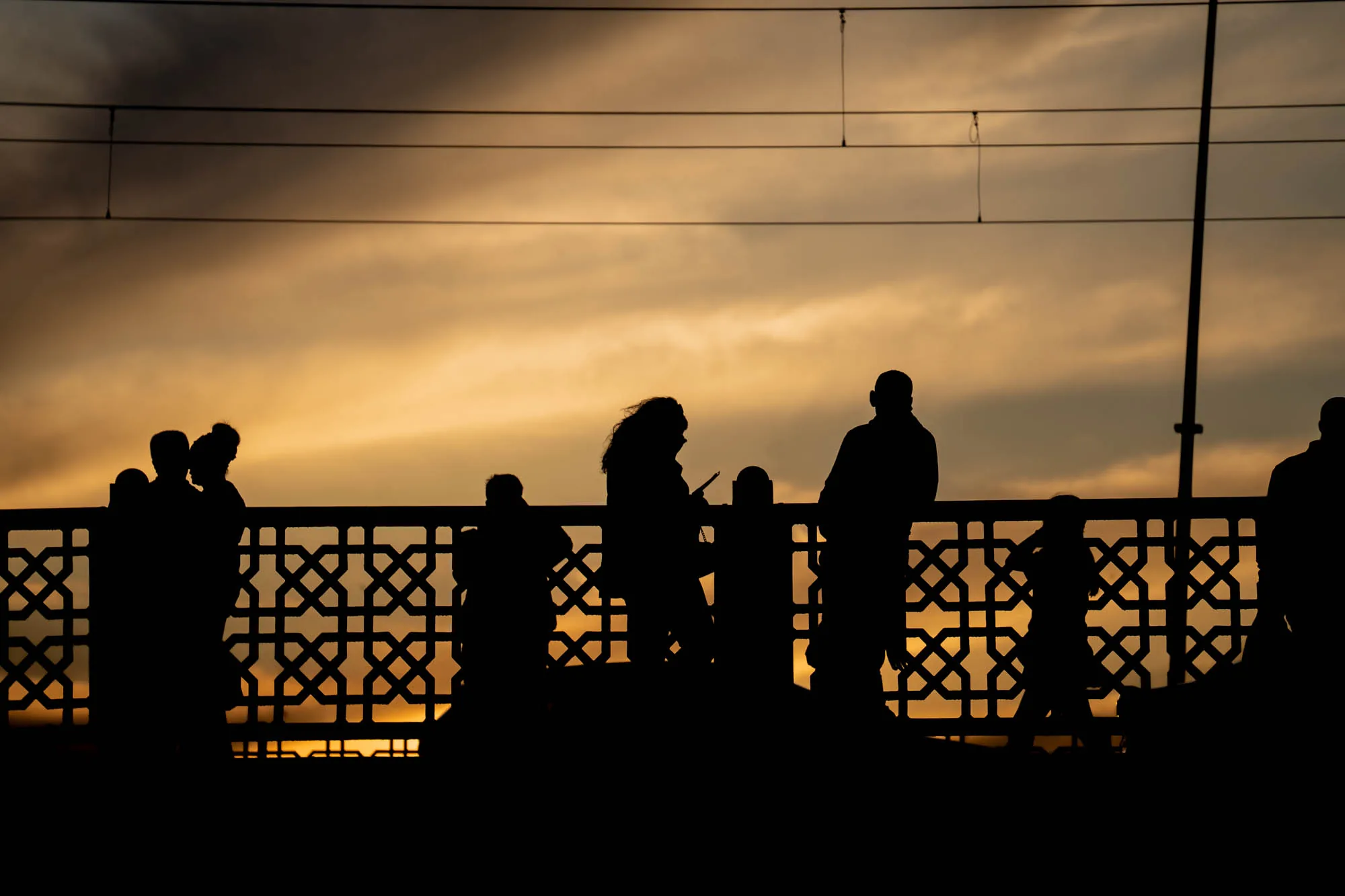 写真はトルコのイスタンブールで撮影された、夕暮れ時のシルエット写真です。画面全体を覆う空は、オレンジ色から暗い灰色へとグラデーションを描き、その美しい夕焼けを背景に、橋の手すりと思われる装飾的な柵の上に立つ人々のシルエットが浮かび上がっています。人々は家族連れやカップルなど、様々なグループで集まっており、風景を眺めたり、スマートフォンを操作したり、それぞれが夕暮れの時間を楽しんでいるようです。手すりの幾何学的な模様と人々のシルエットが組み合わさり、どこかノスタルジックで静寂な雰囲気を醸し出しています。頭上には電線が走り、都市の喧騒をほのかに感じさせます。全体として、イスタンブールの美しい夕暮れと、そこに集う人々の日常を切り取った、印象的な一枚です。