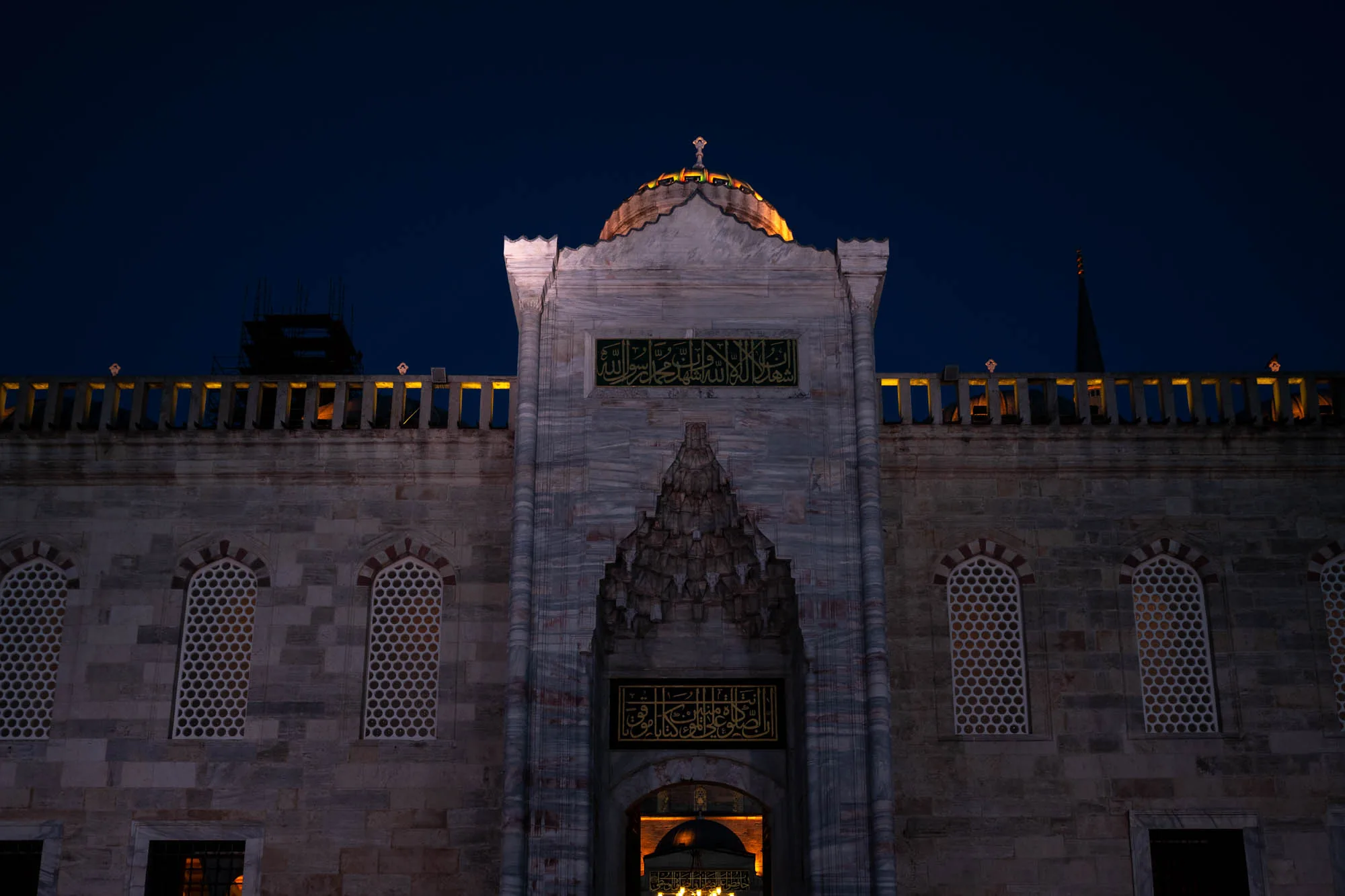 写真はトルコのイスタンブールで撮影されたもので、夜のモスクの正面を捉えています。空は深い青色で、モスクの石造りの壁は薄暗い光に照らされ、灰色がかっています。建物の構造は左右対称で、中央に大きな入り口があり、その上にはドーム型の屋根が載っています。ドームの頂上にはシンボルマークのようなものが確認できます。壁には美しいアラビア文字が刻まれており、イスラム教の雰囲気を醸し出しています。モスクの壁には、格子状の窓が並んでおり、内部の光がほのかに漏れて、幻想的な雰囲気を演出しています。手前には建物の壁に沿って手すりが見え、その背後には工事現場のような足場が確認できます。全体的に静かで神秘的な印象を与える写真です。