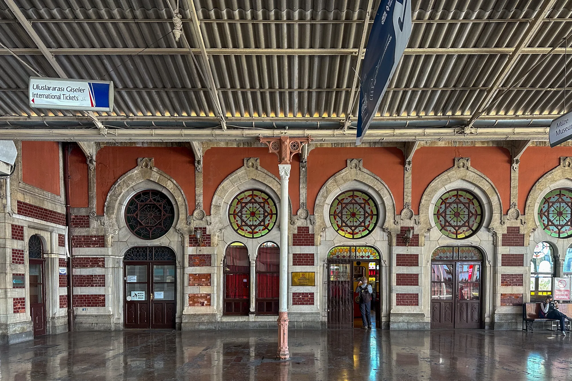 写真はトルコのイスタンブールにある駅の内部を捉えたものです。床は磨かれたように反射しており、天井は金属製の波板で覆われています。壁はレンガと石造りで、アーチ状の窓やドアが並んでいます。窓には幾何学的な模様のステンドグラスがはめ込まれており、壁の色はオレンジ色です。チケット売り場を示す看板や博物館の案内板が見えます。一人の男性が駅構内に入ろうとしており、ベンチに座ってスマートフォンを操作している人もいます。全体的に、歴史的建造物である駅の荘厳な雰囲気を伝えています。