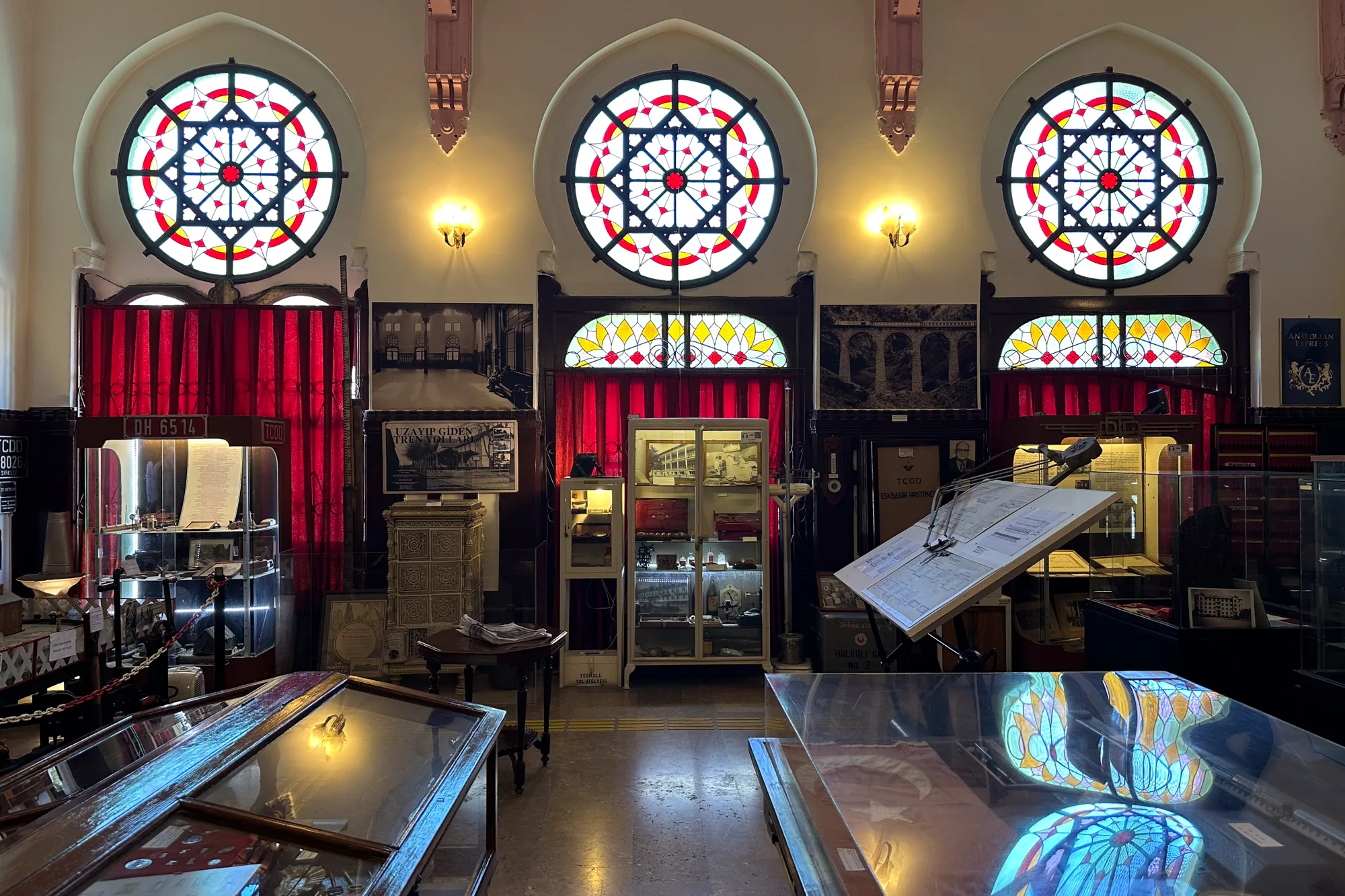 写真はトルコのイスタンブールにある博物館の内部を写したものです。部屋は、鮮やかなステンドグラスの窓、展示ケース、アンティーク家具で飾られています。  3つの大きな円形のステンドグラスの窓が並び、赤、白、黒、黄色の模様が特徴的です。これらの窓の下には、赤いベルベットのカーテンが下がっています。壁には古風な壁掛け照明が設置され、暖かく照らしています。窓の間には、歴史的な写真や文書が飾られています。  部屋には、鉄道関連の品々を展示するガラスケースがいくつかあります。これらのケースの中には、書類、模型、その他鉄道の歴史に関連する様々な遺物が展示されています。床には黄色の点字ブロックが見えます。手前には、ステンドグラスの窓の模様が映り込んでいるガラスケースがあります。全体的に、この写真は歴史的、文化的遺産を保存し展示している博物館の落ち着いた雰囲気を伝えています。