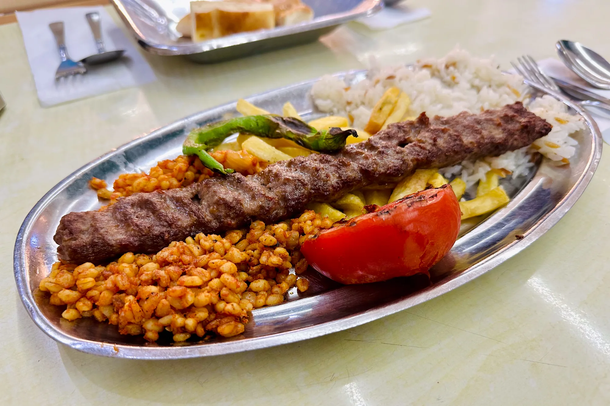 写真はトルコのイスタンブールで撮影された料理の写真です。銀色のトレイに盛り付けられたケバブ定食がメインに写っています。メインディッシュは、細長い形状のケバブで、表面は香ばしく焼かれています。ケバブの横には、白米、フライドポテト、焼いたトマト、焼いたピーマン、麦のような粒状のオレンジ色の料理が添えられています。全体的にボリュームがあり、美味しそうな雰囲気です。背景には、パンが乗ったお盆やカトラリーが写っており、レストランで食事をしている様子が伺えます。テーブルの表面は明るい色で、清潔感があります。