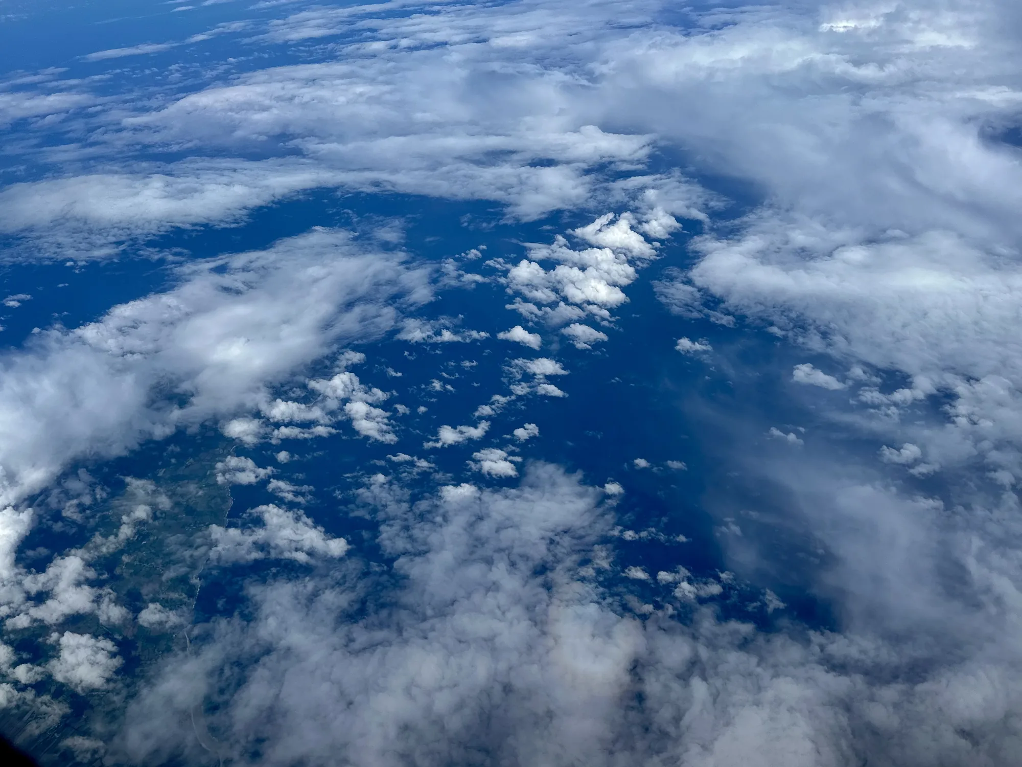 この写真はイギリスのロンドン上空で撮影されたもので、空を覆う雲の様子を捉えています。写真全体は青空と様々な形の白い雲で構成されており、まるで空に浮かぶ白い島々のように見えます。一部には地上の風景も見え、緑豊かな土地が広がっていることが分かります。雲はふんわりとした綿菓子のようなものから、層状に広がるものまで多様な表情を見せており、空の広大さと美しさを感じさせる一枚です。