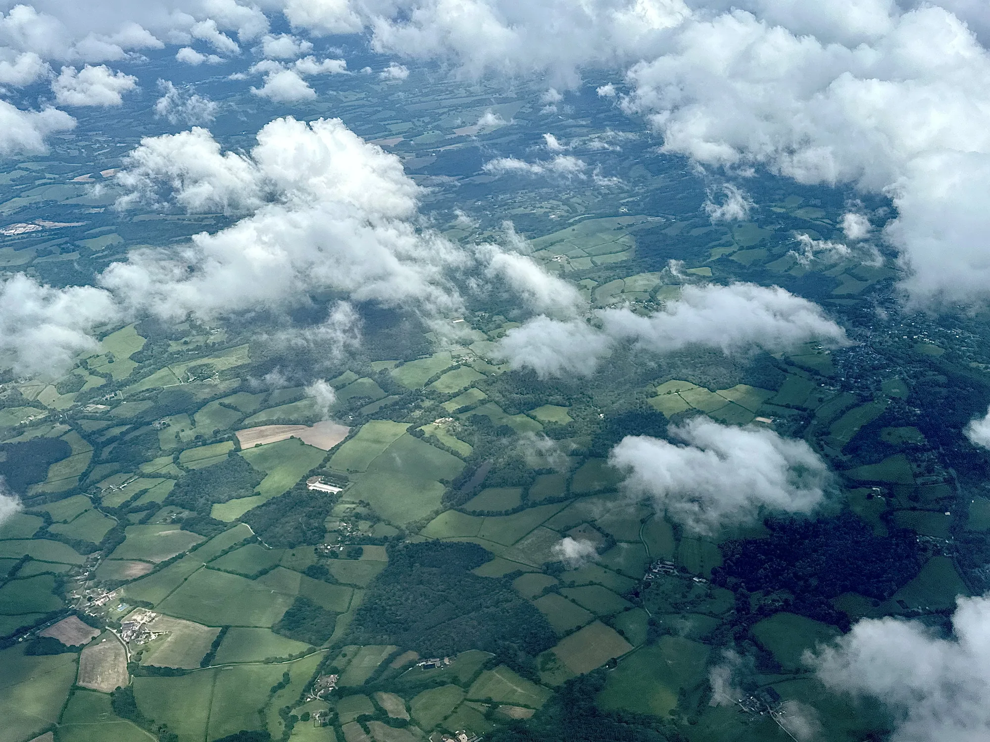 写真は、イギリス、ロンドンの上空から撮影されたと思われる風景を捉えています。視点は高く、まるで飛行機の中から見下ろしているようです。眼下には、パッチワークのように広がる緑豊かな田園地帯が広がっています。畑は様々な緑色に染まり、その境界線は生垣や木々によって区切られています。畑の合間には、森や小さな集落も点在し、自然と人間の営みが調和した風景を作り出しています。画面の上半分は雲に覆われており、太陽の光を浴びて白く輝いています。雲は立体感があり、風景に奥行きを与えています。全体的に、穏やかで平和な雰囲気が漂う写真です。