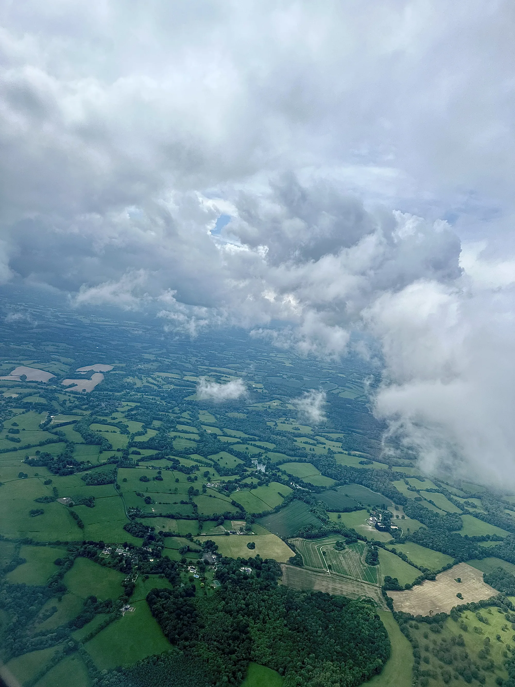 写真は飛行機から撮影されたイギリス・ロンドン郊外の田園風景です。画面の上部は厚い雲に覆われており、ところどころに青空が見えています。雲の下には、緑色の牧草地や畑がパッチワークのように広がっています。牧草地は木々や生垣で区切られ、点在する村や農家の建物が見られます。田園地帯は丘陵地帯であり、遠くまで続く緑のグラデーションが美しいです。湿った空気とあいまって、全体的に落ち着いた、穏やかな雰囲気の写真です。