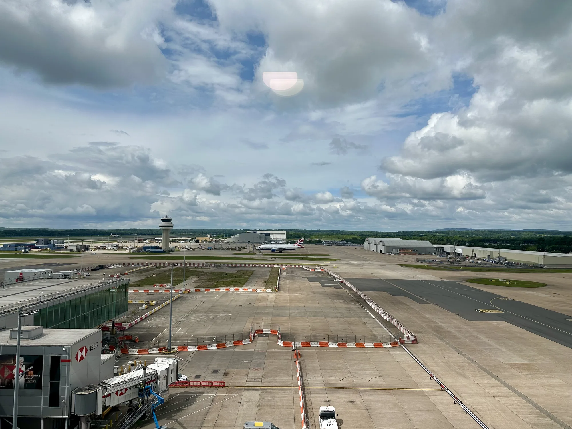 写真は、イギリス・ロンドンの空港の様子を捉えたものです。高い場所から撮影されたようで、空港の滑走路や駐機場、ターミナルビルなどが一望できます。空には白い雲が広がり、晴れ間も見えます。手前にはHSBCのロゴが入った搭乗ブリッジが見えます。滑走路にはブリティッシュエアウェイズの航空機が駐機しており、遠くには管制塔も見えます。全体的に、空港の活気ある雰囲気と、旅行への期待感を感じさせる写真です。