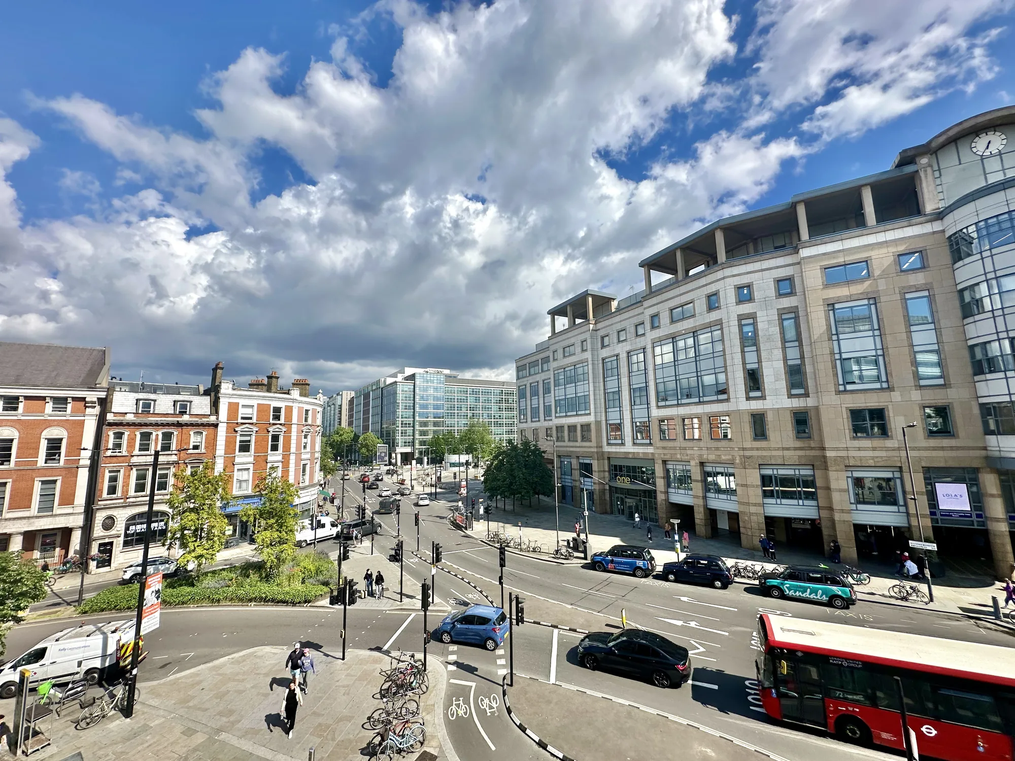 写真はイギリス・ロンドンの街並みを上から見下ろした風景です。空には白い雲が広がり、晴れた日のようです。道路には車やバスが走り、歩道には人が行き交っています。赤レンガの建物や近代的なオフィスビルなど、様々な建築様式が混在しており、ロンドンの多様な表情が感じられます。道路標識や自転車専用レーンなども確認でき、街の機能性がうかがえます。赤いロンドンバスが特徴的で、都市の活気ある雰囲気を感じさせる一枚です。