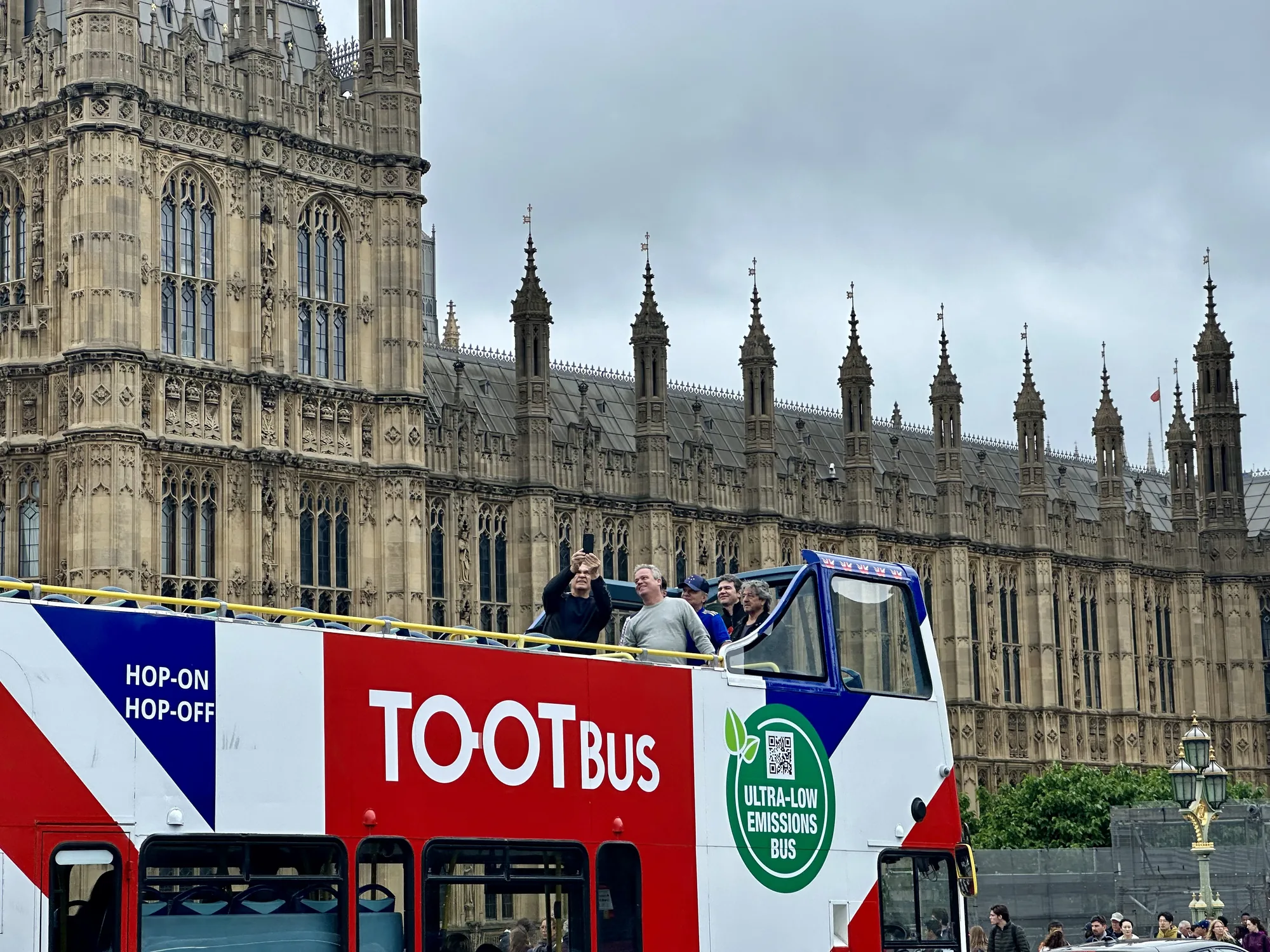ロンドンの国会議事堂を背景に、観光客を乗せたオープントップの二階建てバスが写っています。バスは赤、白、青のイギリス国旗を模したカラーリングで、「TOOT BUS」と大きく書かれています。バスの上には観光客が数人おり、景色を眺めたり、写真を撮ったりしているようです。背景の国会議事堂は、ゴシック様式の荘厳な建物で、細部まで丁寧に装飾されています。空は曇っていて、全体的に落ち着いた雰囲気の写真です。バスには「HOP-ON HOP-OFF」や「ULTRA-LOW EMISSIONS BUS」という文字が見えます。