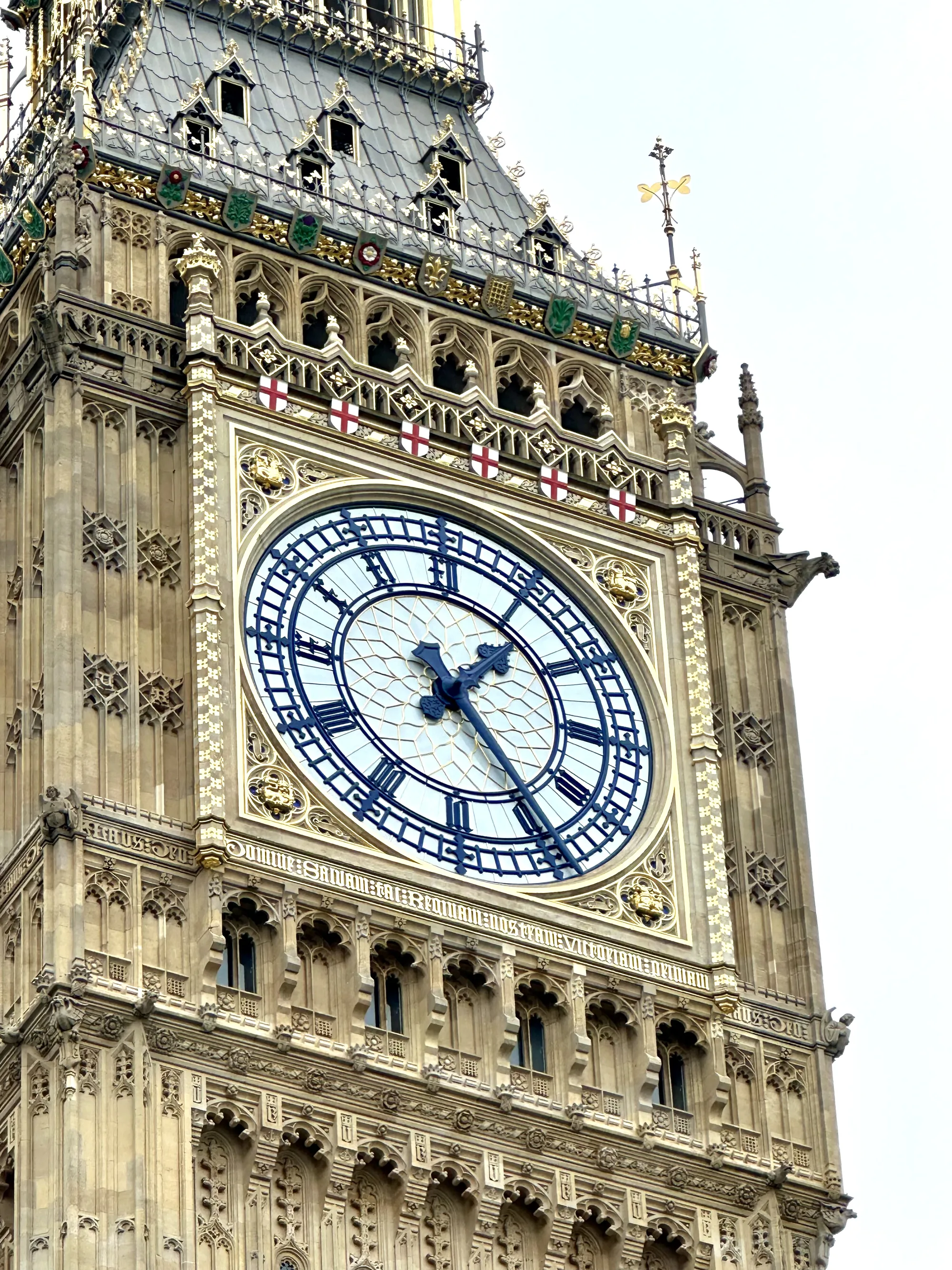 この写真は、イギリスのロンドンにあるウェストミンスター宮殿の時計台、通称ビッグ・ベンの下部を捉えています。画面の中心には、有名な時計の文字盤が大きく配置されており、濃い青色のローマ数字と針が印象的です。文字盤のデザインは緻密で、歴史を感じさせる風格があります。時計台の石造りの壁面は、細部にわたる装飾が施されており、ゴシック建築の荘厳さを伝えています。壁面には窓がいくつか見られ、上部には紋章や彫刻が飾られています。空は白く霞んでおり、建物の重厚感を際立たせています。全体的に、歴史的建造物の美しさと、ロンドンの象徴的な風景を捉えた写真と言えるでしょう。