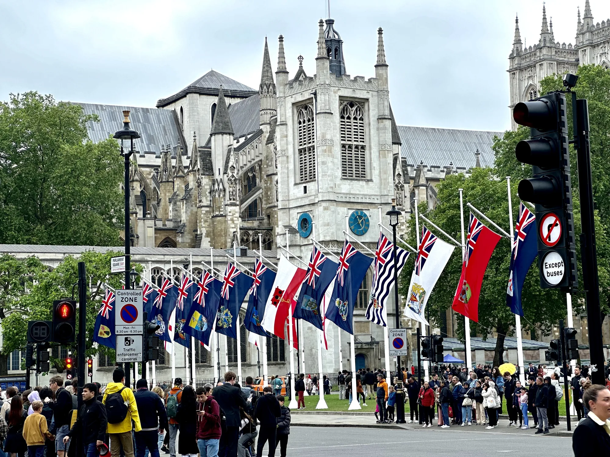 写真はイギリスのロンドンで撮影されたもので、ウェストミンスター寺院を背景に、多くの旗が掲げられた風景が写っています。手前には横断歩道があり、信号機は赤で人が立ち止まっています。多くの人々が寺院の方を向いており、観光客や地元の人々が集まっているようです。旗はイギリスの国旗であるユニオンジャックをベースにしたデザインで、各国の紋章や図柄が入っているようです。空は少し曇っており、落ち着いた雰囲気です。交通標識には「Controlled Zone」と書かれています。