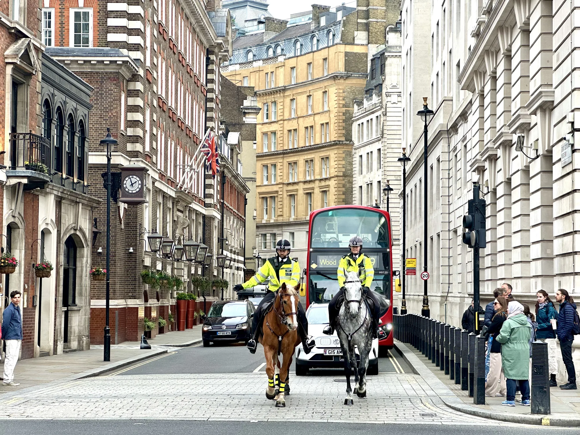 写真はイギリスのロンドンで撮影された街並みを捉えています。石畳の道路の中央には、騎馬警官が2人、それぞれ茶色と灰色の馬に乗り、こちらに向かって歩いています。2人の警官は蛍光色の制服を着用しており、ヘルメットを被っています。彼らの背後には赤い二階建てバスが走り、さらに奥には自動車が走行しています。道路の両脇には歴史を感じさせるレンガ造りや石造りの建物が立ち並び、その中にはユニオンジャックが掲げられている建物もあります。左側の建物には時計が設置され、植木鉢で飾られた窓が見られます。右側の歩道には数人の人々が立っており、街の様子を眺めているようです。全体的に落ち着いた雰囲気で、ロンドンの日常風景が切り取られています。