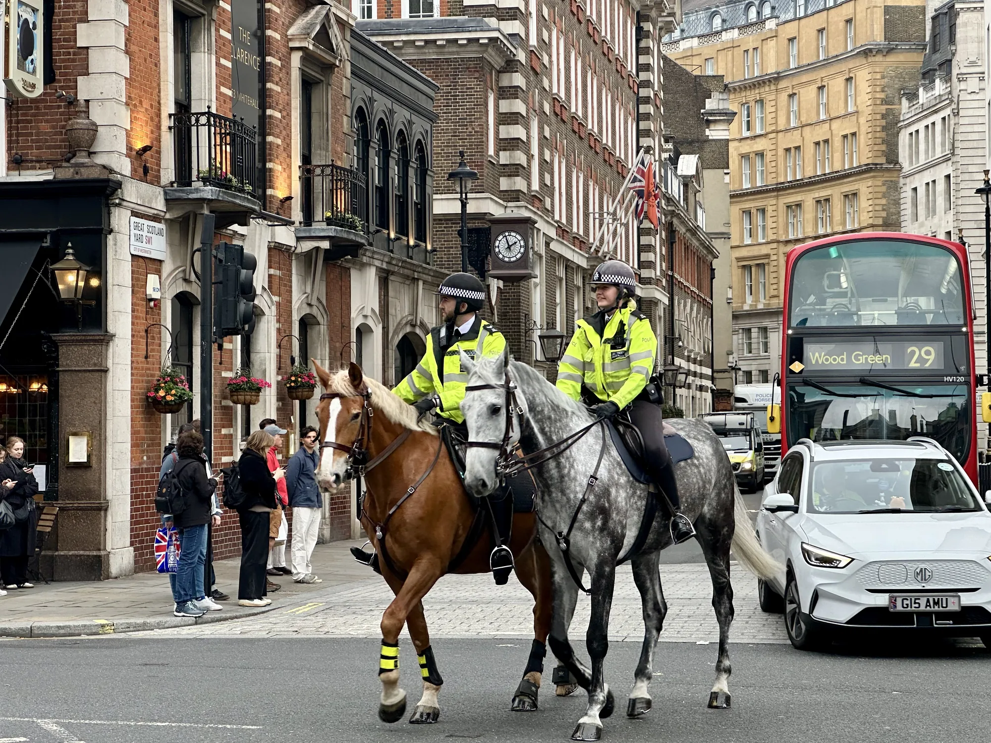 写真はイギリスのロンドンで撮影されたと思われるもので、市街地の道路を騎馬警官が横断している様子が捉えられています。2名の警官がそれぞれ馬に乗っており、画面奥に向かって進んでいます。警官は蛍光色のジャケットを着用し、ヘルメットを被っています。手前の馬は茶色で、後ろの馬は灰色です。  背景には赤レンガ造りの建物が並び、そのうちの一つには「The Clarence」という看板が見えます。また、「Great Scotland Yard SW1」という道路標識も確認できます。建物の上にはイギリス国旗が掲げられています。道路には赤い二階建てバスや白い車など、複数の車両が走行しています。歩道には数人の人々が立っており、スマートフォンを見ている人もいます。曇り空で、全体的に落ち着いた雰囲気の写真です。