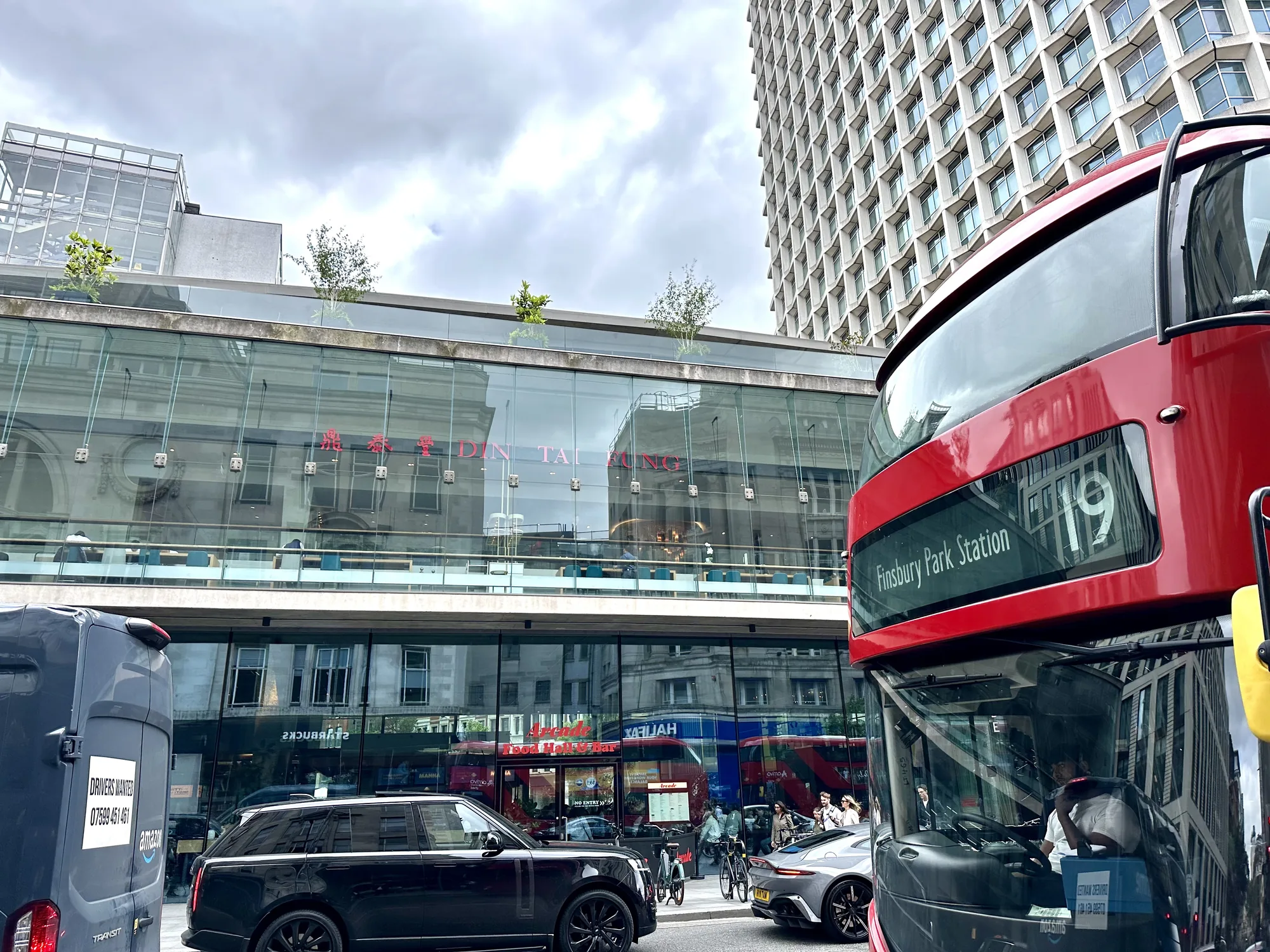 写真は、イギリスのロンドンの街並みを捉えています。手前には鮮やかな赤い二階建てバスの一部が写っており、「19 Finsbury Park Station」と書かれた行先表示が見えます。バスの運転手も確認できます。バスの向こうには、ガラス張りの建物が見え、そこには「DIN TAI FUNG」という赤い文字の看板が掲げられています。建物内にはテーブルと椅子が並び、人が座っている様子が伺えます。また、建物の上部には緑の木々が植えられており、都会の中の緑を感じさせます。  写真には他にも、黒い高級車や白い車、配達用のバンなどが写っており、活気のある街の様子が伝わってきます。建物のガラスには周囲の建物や空が反射し、複雑な光景を作り出しています。空は曇り空で、落ち着いた雰囲気を醸し出しています。全体的に、ロンドンの都会的な風景と人々の生活が感じられる写真です。配達バンの車体には「DRIVERS WANTED 07599 451 461」と書かれた求人広告が見えます。