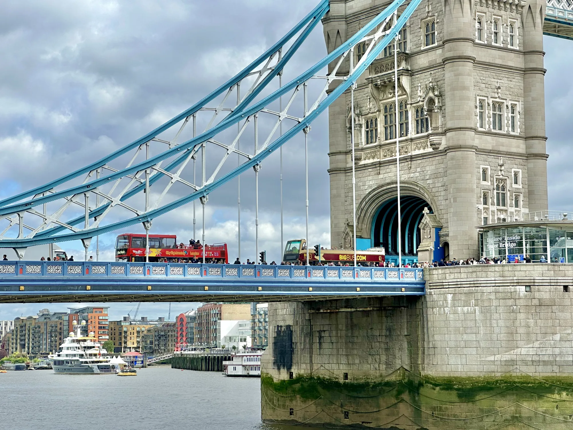 写真には、イギリス・ロンドンの象徴的な建造物であるタワーブリッジが写っています。空は曇り空で、橋の青い鉄骨が印象的です。橋の上には、観光客を乗せた2階建てバスや、多くの人々が歩いているのが見えます。橋の塔部分は美しい石造りで、細部まで丁寧に装飾されているのがわかります。橋の下にはテムズ川が流れ、豪華なヨットや観光船が浮かんでいます。写真全体から、ロンドンの歴史と活気が感じられます。