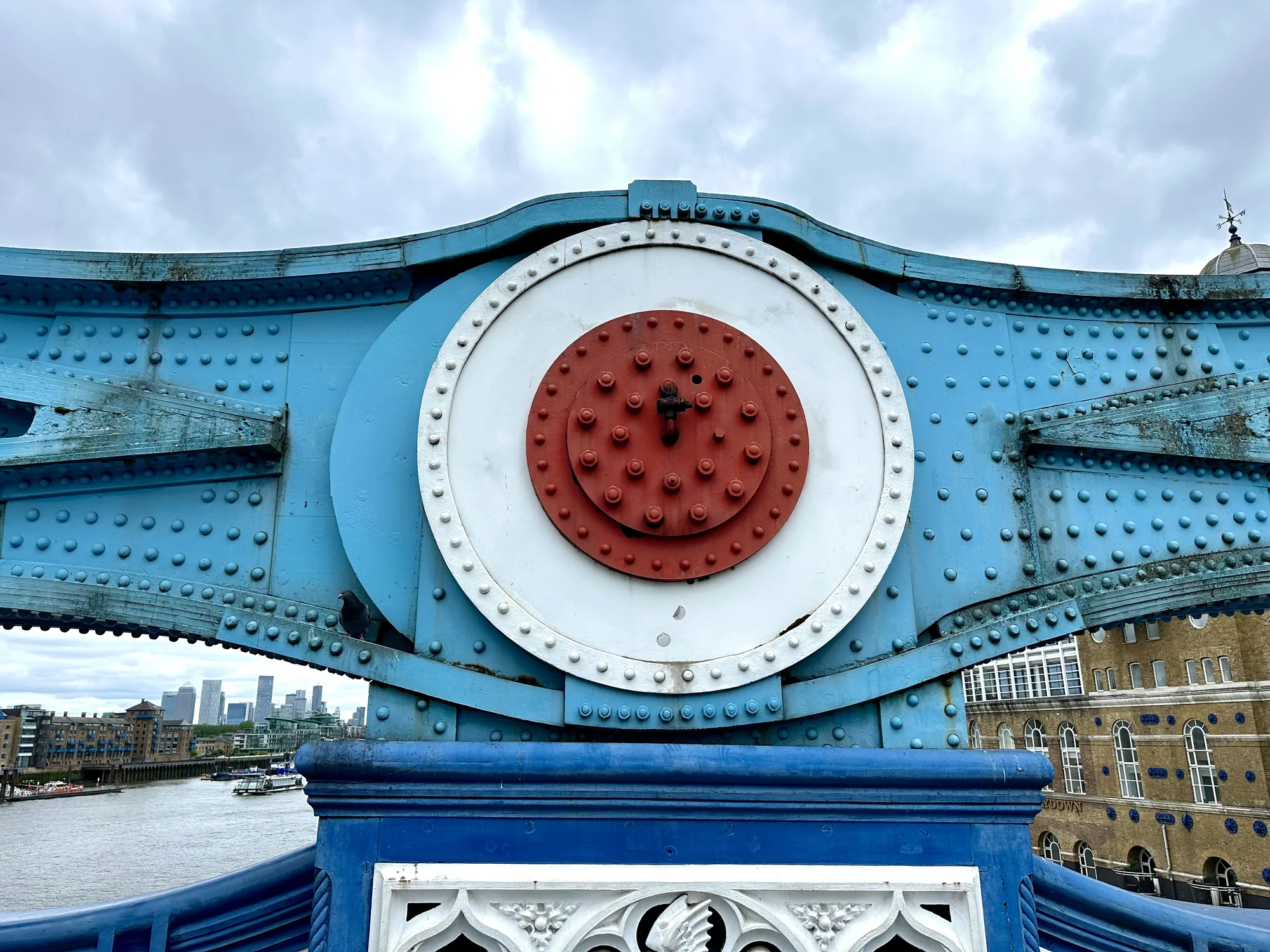 写真は、ロンドンのタワーブリッジの一部を撮影したものです。橋の構造物である、青色の鉄骨とリベットが印象的で、中央には赤、白、青のターゲットのような装飾が施されています。背景には、ロンドンの街並みとテムズ川が見え、曇り空が広がっています。橋の装飾的な手すりも写っており、橋の歴史的な雰囲気を伝えています。全体的に、橋の構造美とロンドンの風景が組み合わさった、興味深い写真です。