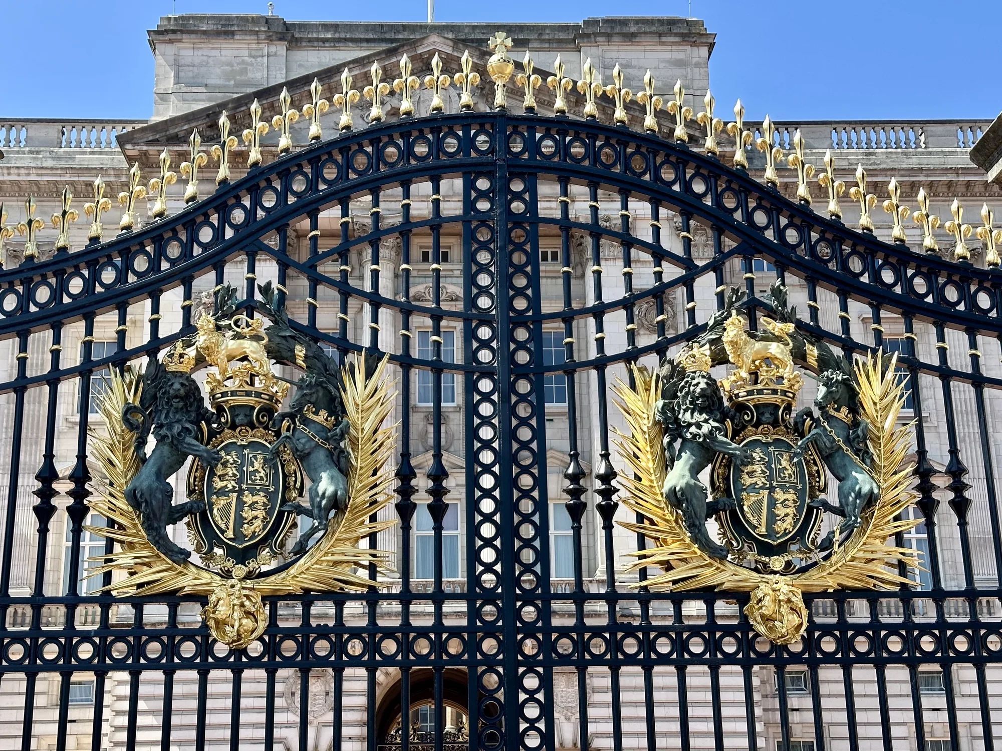 写真はイギリス、ロンドンのバッキンガム宮殿の門を捉えたものです。門は黒色の鉄製で、細い縦棒と円形の装飾が規則的に配置されています。門の上部には金色の装飾が施されており、ユリの紋章のようなモチーフが並んでいます。門の中央には、金色のライオンとユニコーンが盾を支える紋章が左右対称に配置されています。これらの紋章は王冠を戴き、金色の装飾が豪華さを際立たせています。背景には、宮殿の一部が見え、石造りの壁と窓が確認できます。空は晴れており、全体的に明るい雰囲気です。写真全体から、バッキンガム宮殿の格式と壮麗さが伝わってきます。