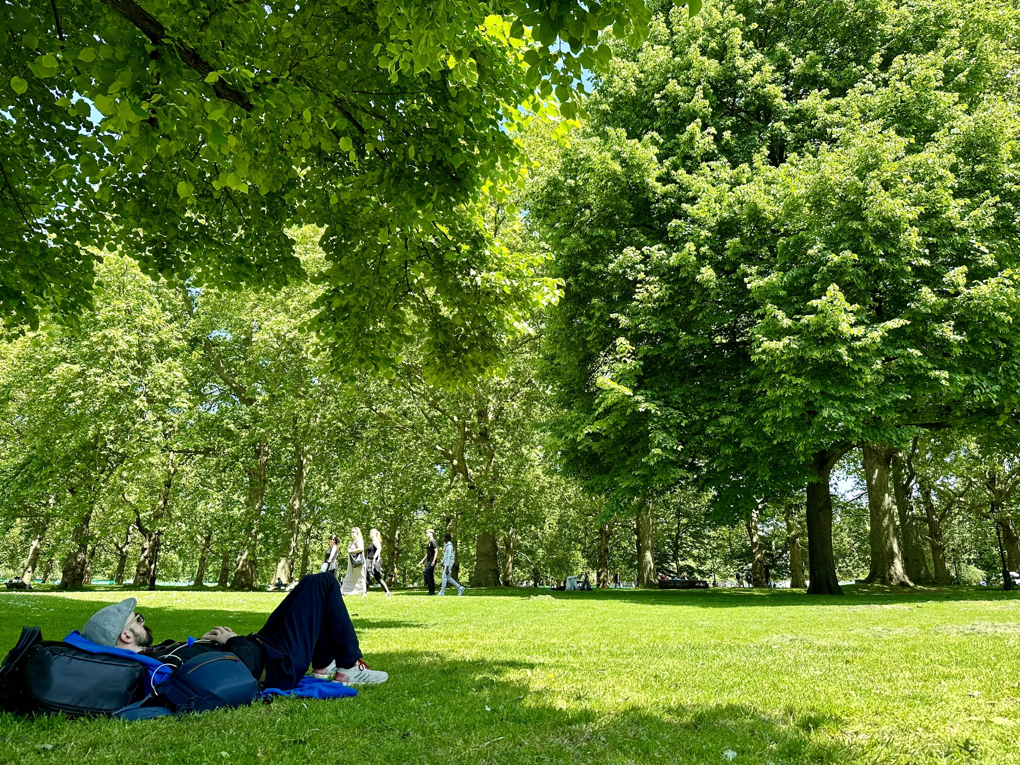 写真は、イギリスのロンドンにある公園の風景を捉えています。前景には、リュックを枕にして芝生の上に寝転んでいる男性がいます。彼は帽子をかぶり、リラックスした様子で空を見上げています。芝生は鮮やかな緑色で、ところどころに木漏れ日が差し込んでいます。背景には、高くそびえる緑豊かな木々が並び、木々の間からは青空がわずかに見えています。木々の間を人々が歩いている様子も確認できます。全体的に、穏やかで平和な雰囲気が漂っており、都会の中のオアシスといった印象を受けます。光の加減や鮮やかな緑色が、写真全体に明るい印象を与えています。