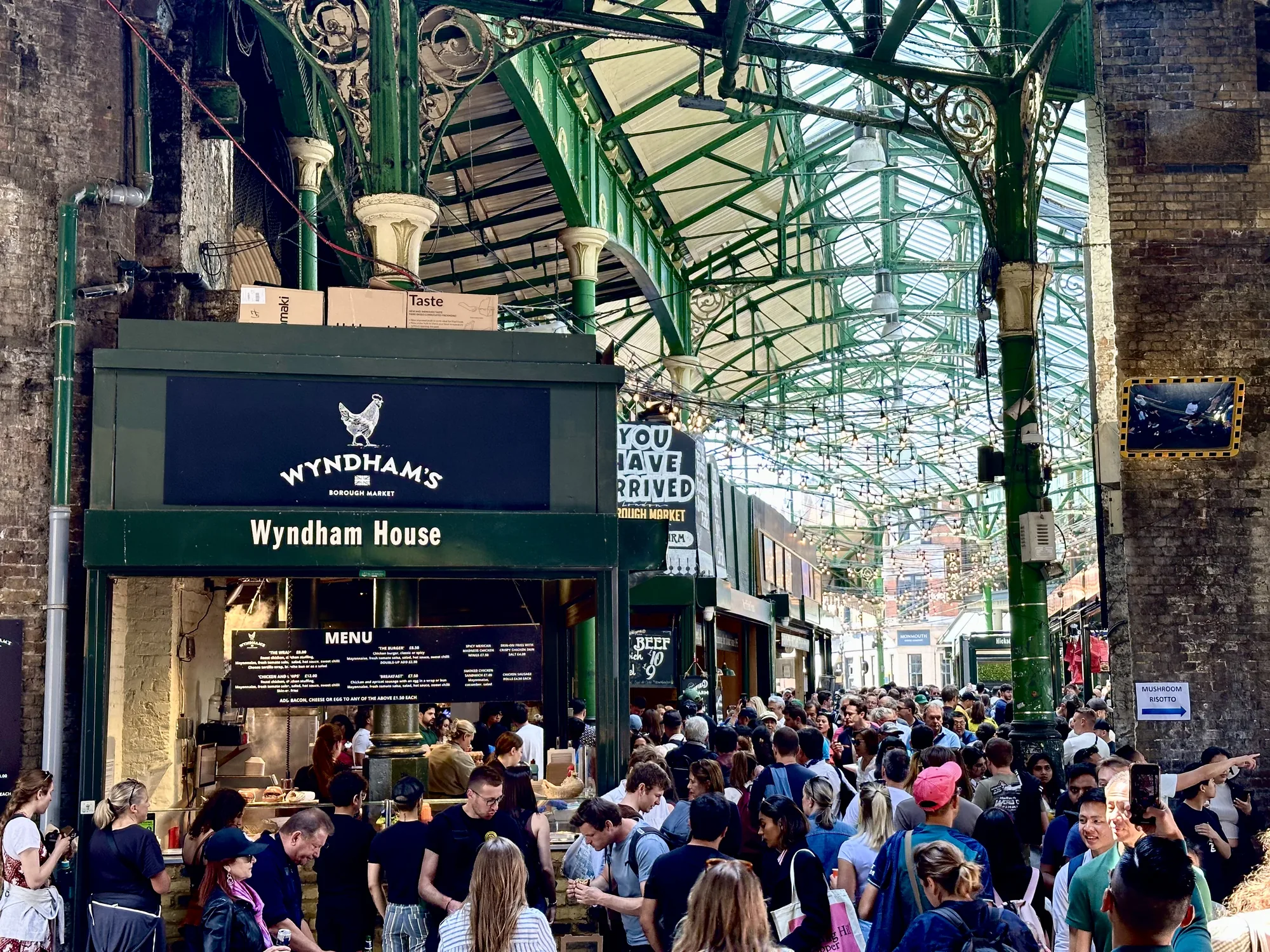 写真はイギリス・ロンドンのマーケットの様子を捉えています。大勢の人々で賑わっており、通路は人で埋め尽くされています。天井は緑色の鉄骨で組まれ、ガラス張りになっており、そこから自然光が差し込んでいます。天井には電球が連なって装飾されています。  写真手前には「WYNDHAM'S BOROUGH MARKET Wyndham House」と書かれた緑色の看板を掲げる店舗があり、メニューが掲示されています。人々が食べ物を求めて列を作っています。レンガ造りの壁には、パイプや配線が走っており、歴史を感じさせる趣です。奥には別の店舗も見え、マーケット全体が活気に満ちている様子が伝わってきます。
