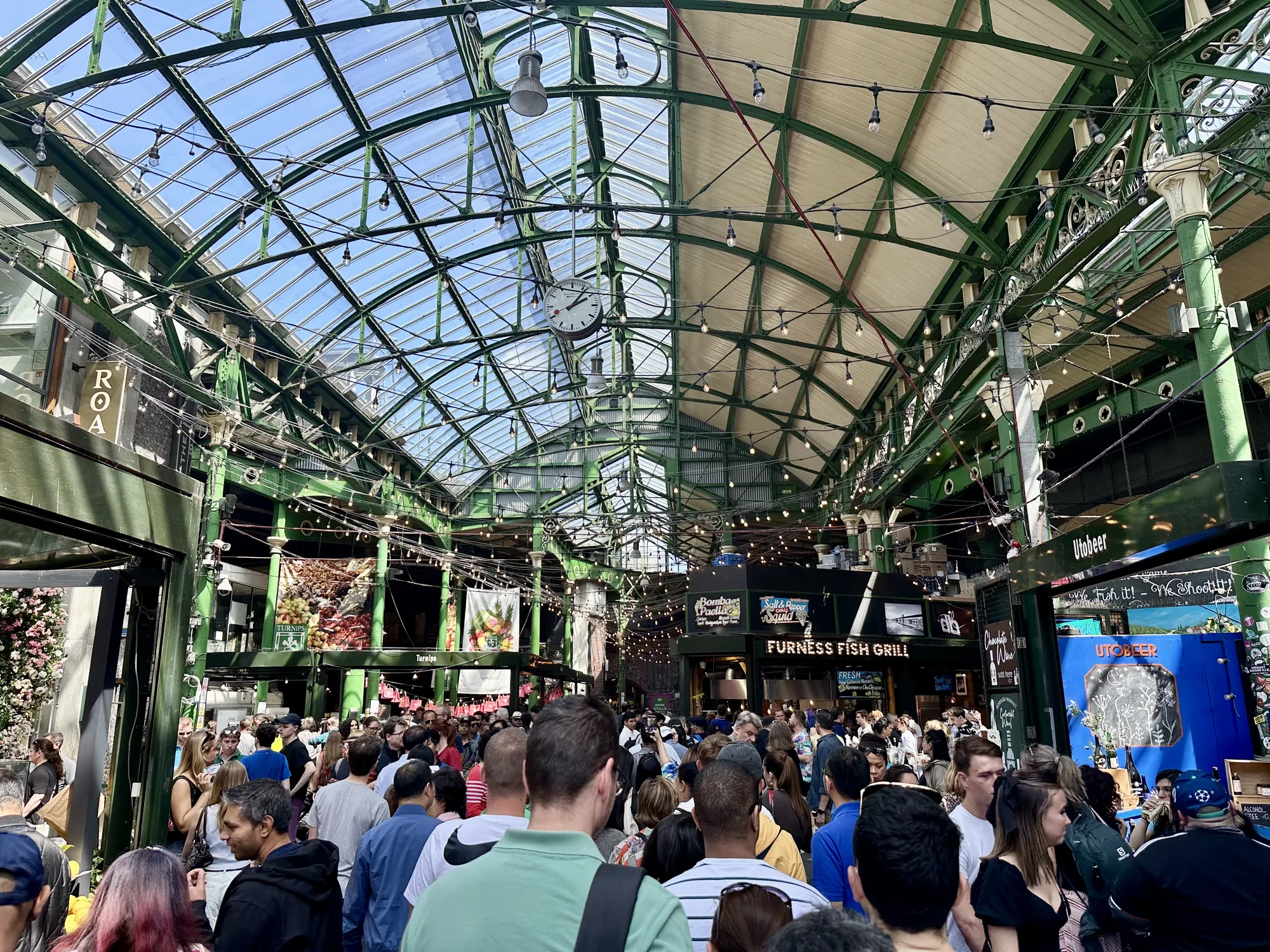 写真は、イギリスのロンドンにある市場の様子を捉えています。ガラス張りの屋根を持つ建物の中は、多くの人で賑わっています。天井は緑色の鉄骨で組まれ、多数の電球が吊り下げられており、独特の雰囲気を作り出しています。  写真には、さまざまな飲食店や店舗が写っています。「FURNESS FISH GRILL」という看板が見え、魚介類を提供する店のようです。他にも、「Utobeer」と書かれた青い看板の店や、野菜や果物を扱う店など、多様な店が並んでいることがわかります。  人々はそれぞれに買い物を楽しんだり、食事をしたりしている様子で、活気に満ちた雰囲気が伝わってきます。写真全体から、ロンドンの市場の賑わいと多様性を感じ取ることができます。