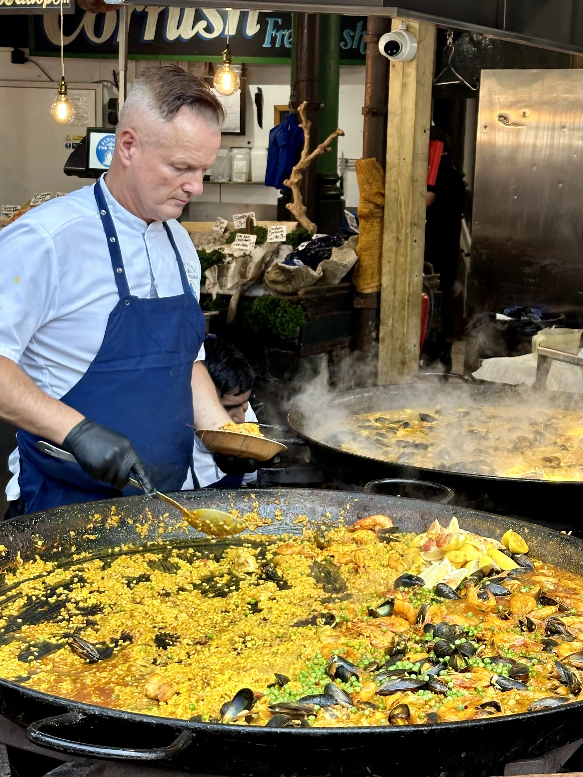 この写真はイギリスのロンドンで撮影されたものです。大きなパエリア鍋が手前にあり、その中には黄色いご飯とムール貝、エビ、グリーンピースなどの具材がたっぷり入っています。鍋は黒く、取っ手が付いています。パエリアを作っている料理人が写っており、白いコックコートに青いエプロンを身につけ、黒い手袋をしています。彼は真剣な表情でパエリアを調理しており、大きなスプーンで混ぜています。背景には別の大きな鍋があり、そこでもパエリアが調理されているようです。蒸気が立ち上り、調理の熱気が伝わってきます。店の看板や照明器具が背景に写っており、市場のような場所で撮影されたことが分かります。活気があり、美味しそうな雰囲気が伝わる写真です。