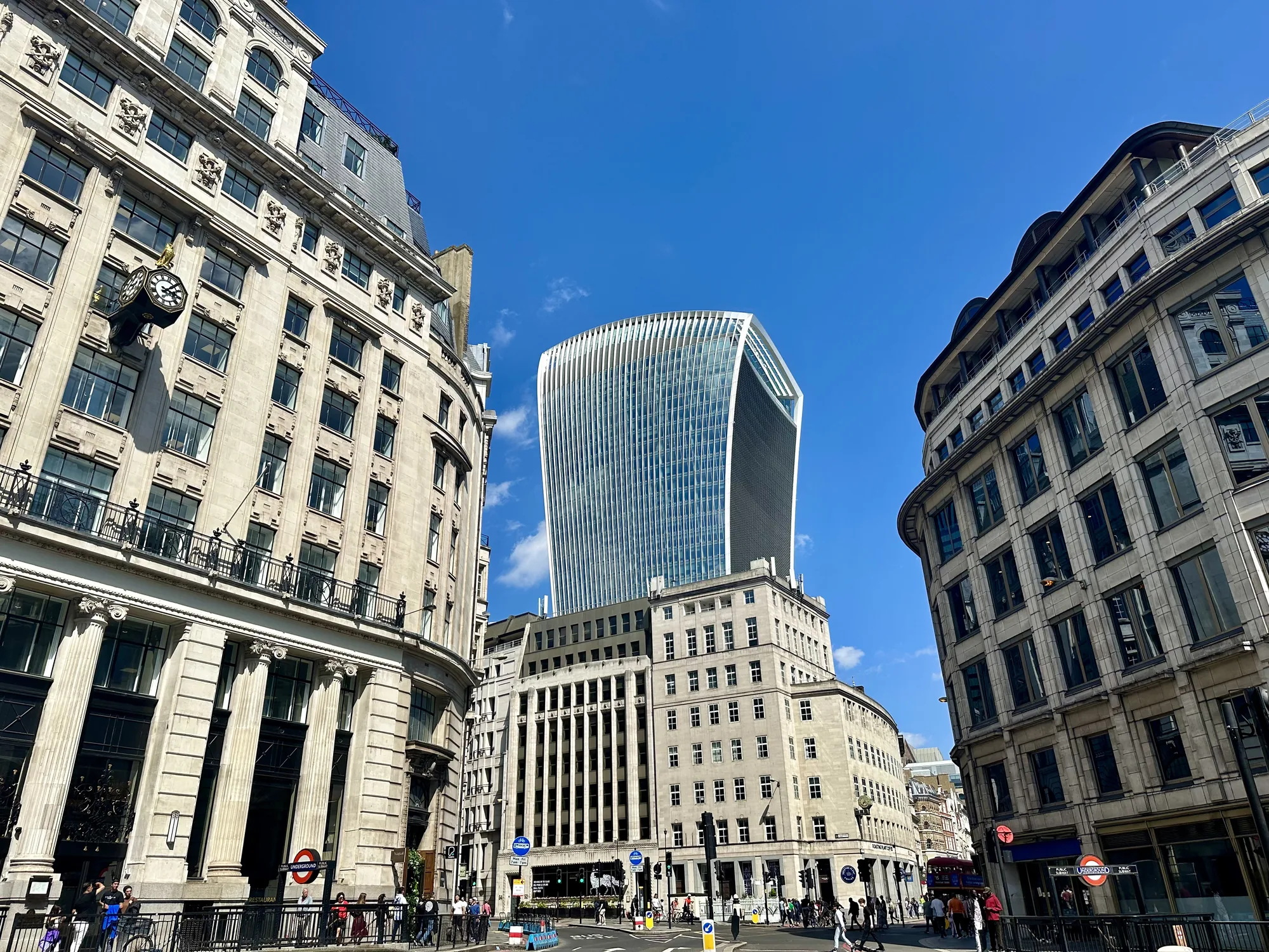 写真はイギリスのロンドンの街並みを撮影したものです。青空を背景に、特徴的な形の高層ビル「ウォークイートーキー（20 Fenchurch Street）」が中央にそびえ立っています。周囲には、歴史を感じさせる石造りの建物が建ち並び、現代的なビルとのコントラストが印象的です。通りの向こう側には地下鉄の標識が見え、人々が行き交っており、ロンドンの日常風景が垣間見えます。暖かく晴れた日の光景で、活気のある街の雰囲気が伝わってきます。